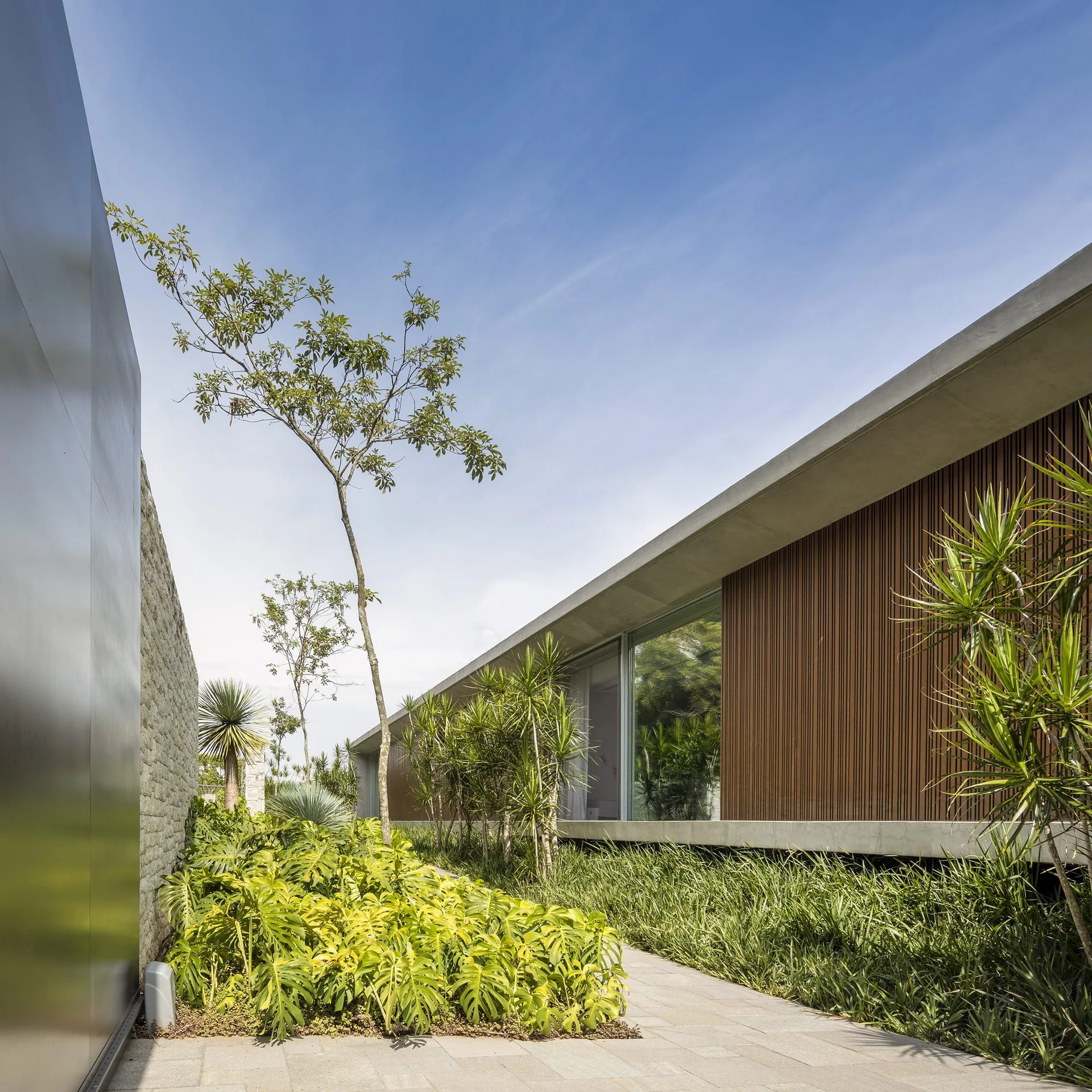A modern concrete house with brown vertical wood slats stands next to a paved stone pathway, lush green garden beds, and a textured masonry wall.