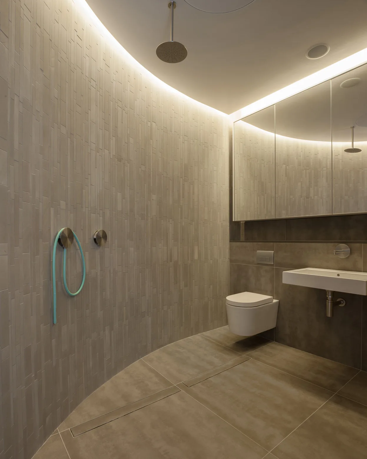 Minimalist wet room featuring vertically stacked tile walls, floating toilet and sink, ceiling-mounted shower head, and bright indirect cove lighting.