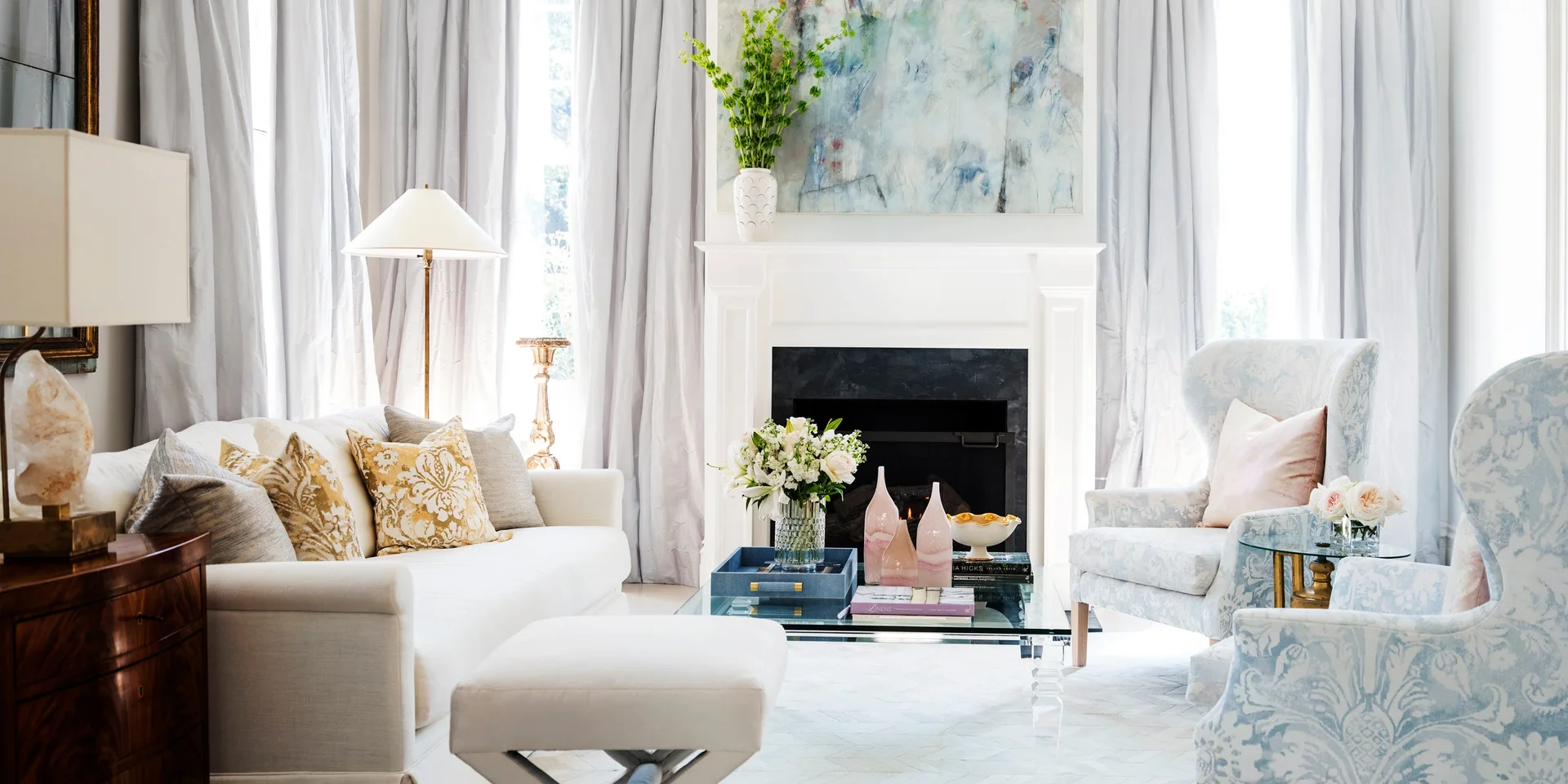 A bright living room features a white fireplace mantel, a cream sofa, and two blue patterned wingback chairs around a glass coffee table.