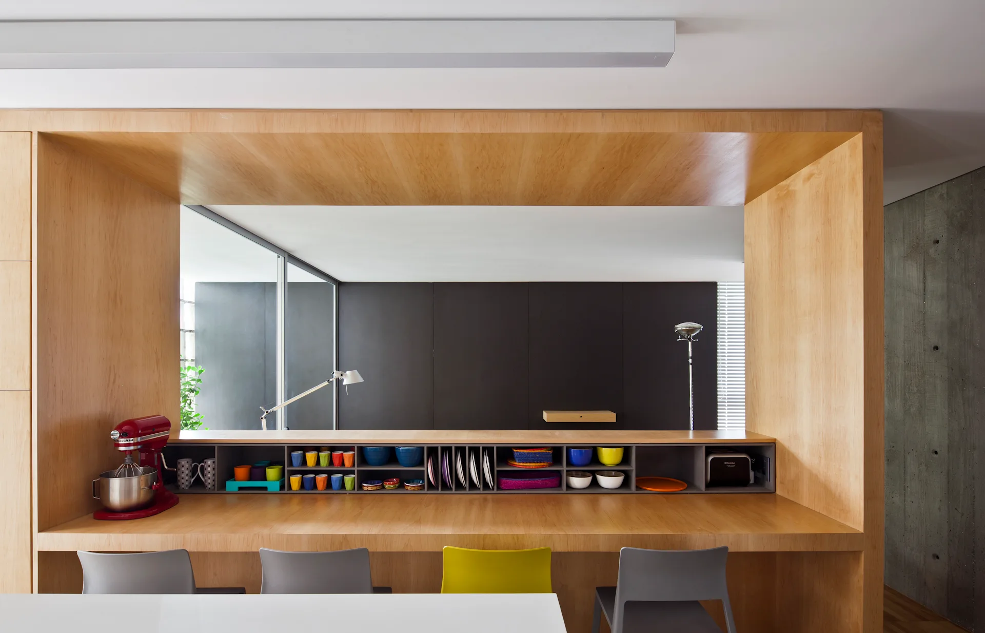 A wood-encased kitchen counter features integrated open shelving filled with colorful dishes and a red stand mixer, with modern dining chairs in front.