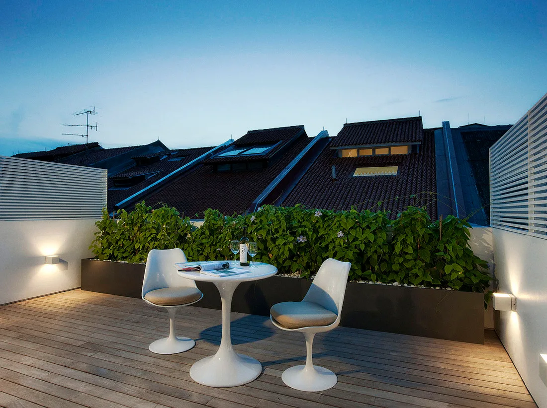 A white pedestal bistro set sits on a wooden deck of a rooftop terrace beside a long planter box and lit wall sconces at dusk.