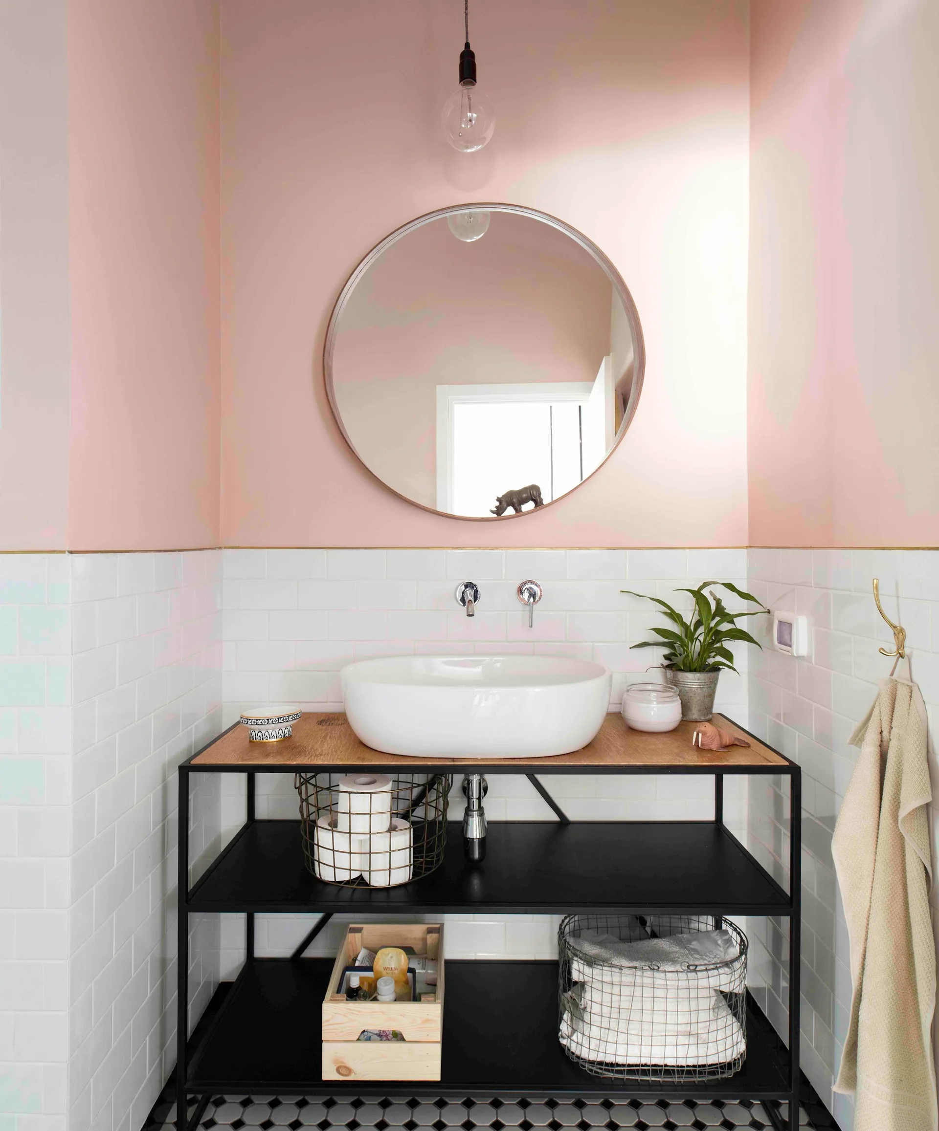A white vessel sink sits on a black metal open-shelf vanity against white subway tiles and soft pink upper walls with a round mirror.