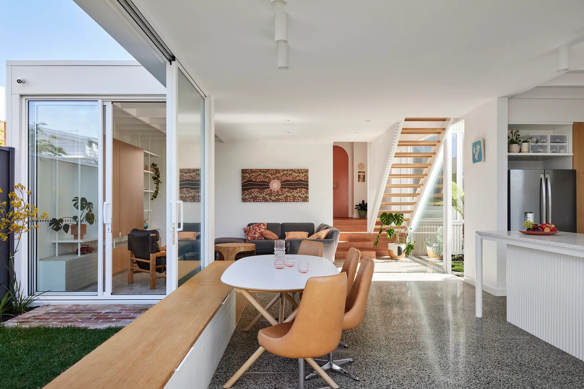 An open-plan dining room features a white oval table with tan leather chairs, a built-in wooden bench, and polished terrazzo flooring.