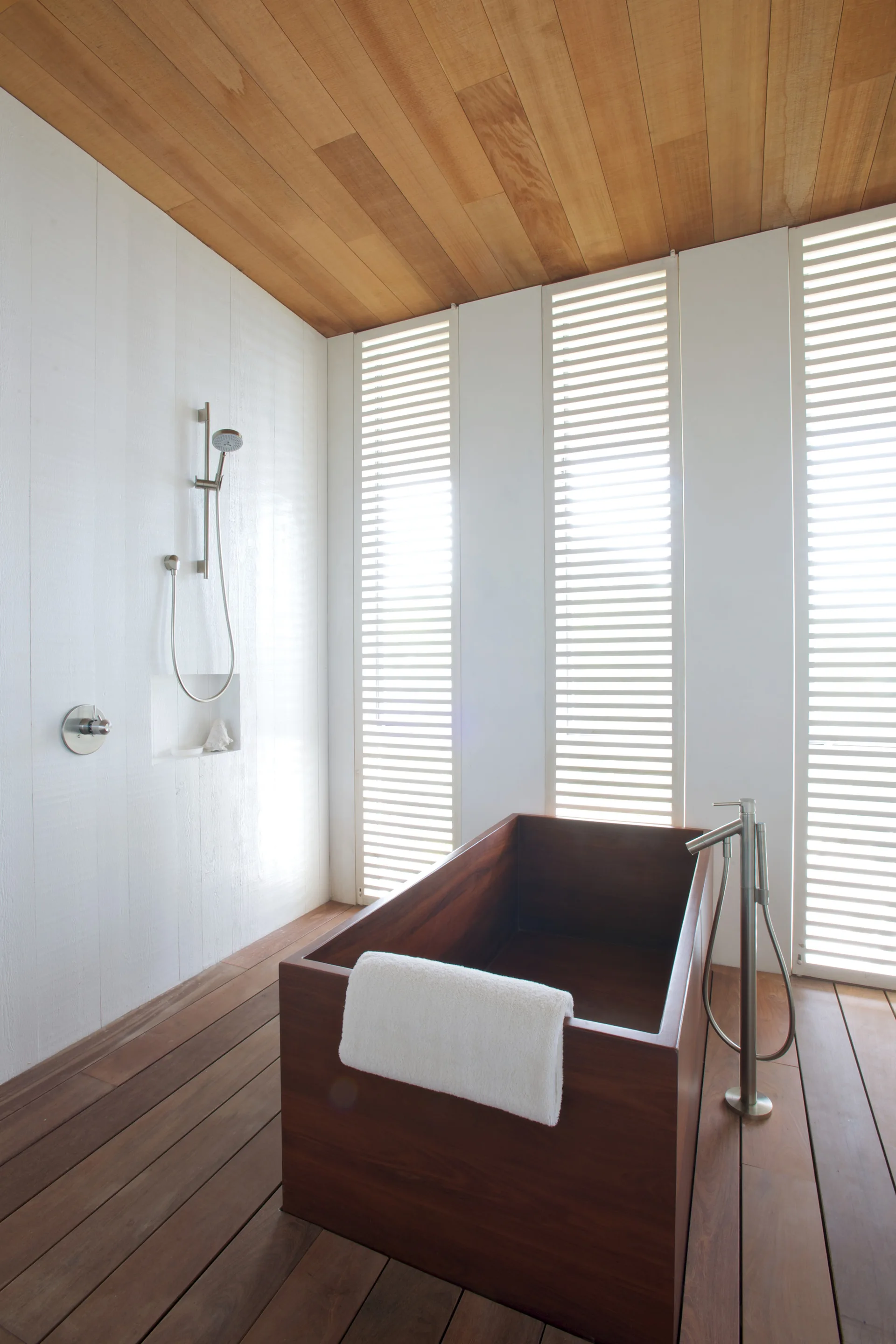 A minimalist bathroom featuring a rectangular wooden soaking tub, matching wood floors and ceiling, and tall white slatted windows.