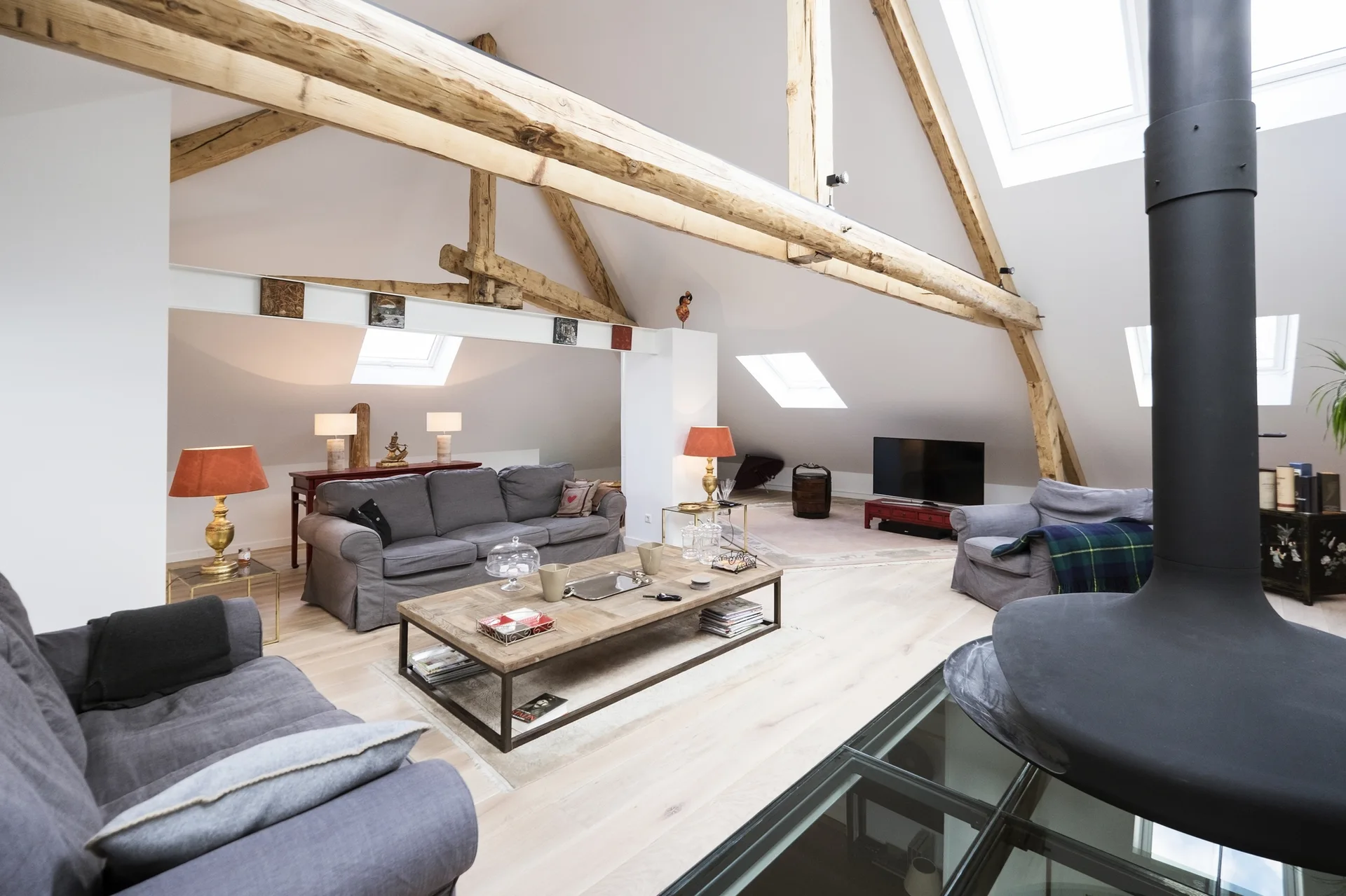 A spacious attic living room with exposed timber beams, grey sofas, a central wood coffee table, and skylights under a pitched ceiling.