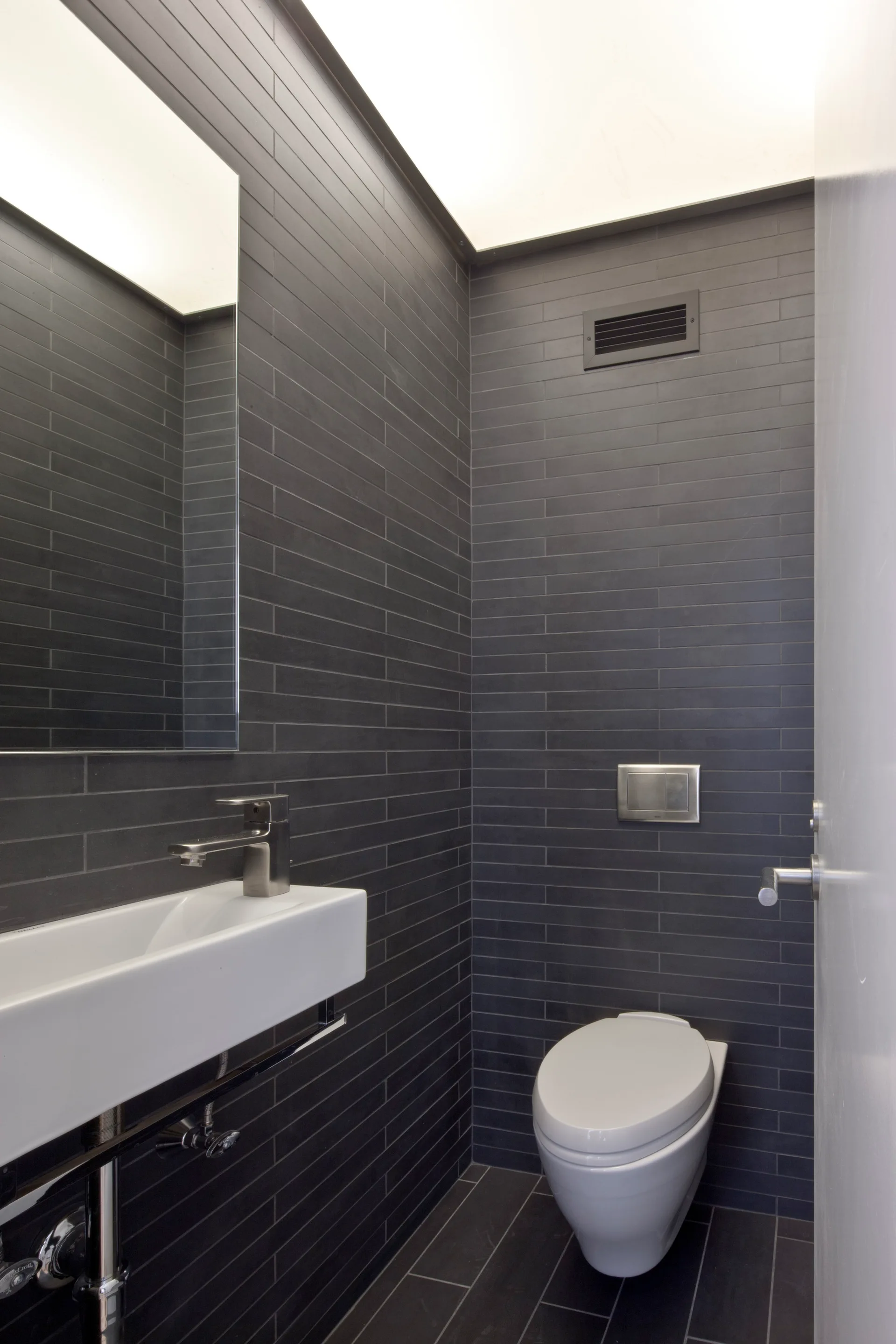 A minimalist powder room featuring dark gray tiled walls and floor, a white floating sink, and a wall-mounted toilet under an illuminated ceiling.