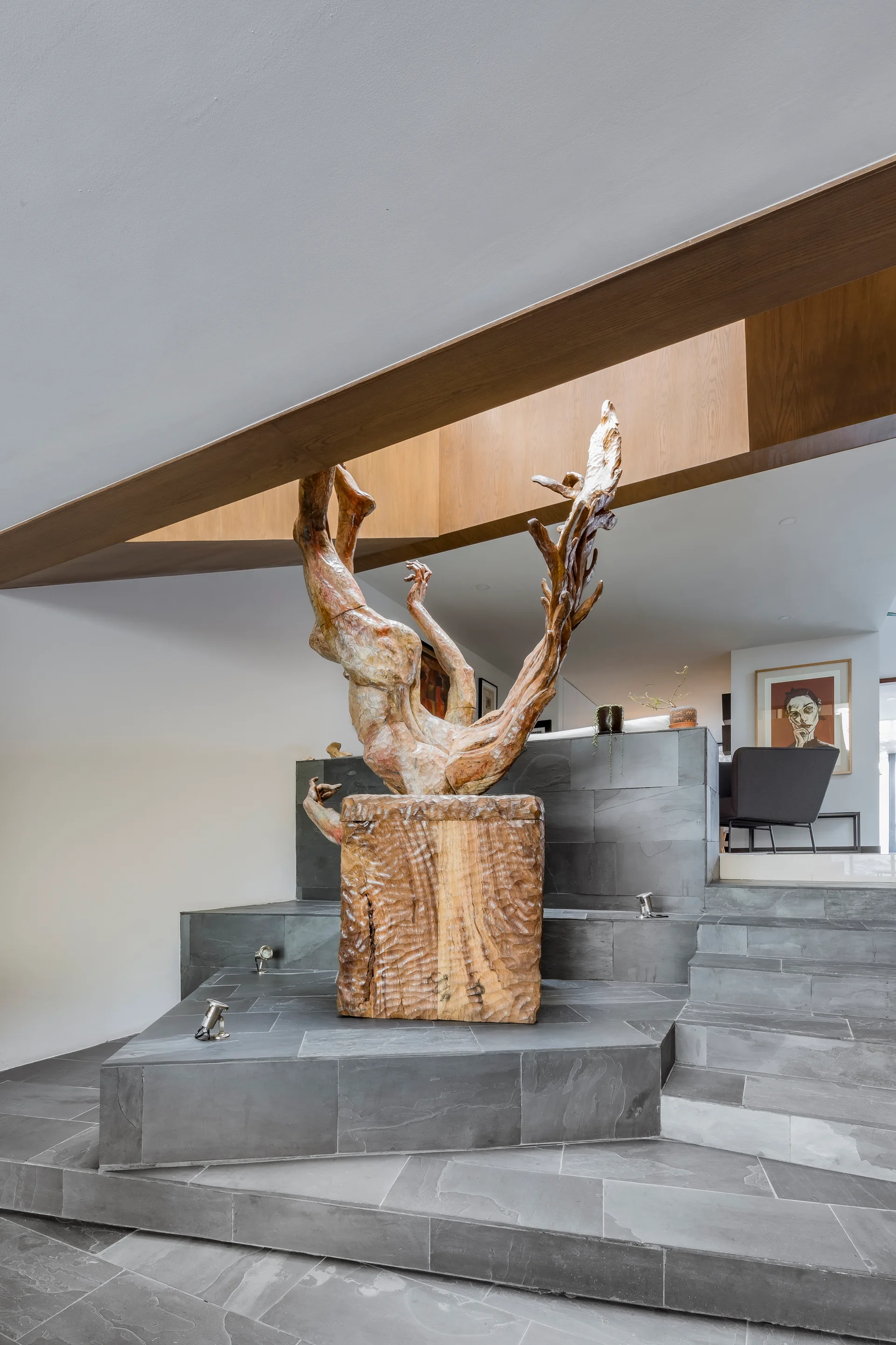 An abstract wooden sculpture stands on a tiered grey slate platform in an entryway with stairs and an angled ceiling.