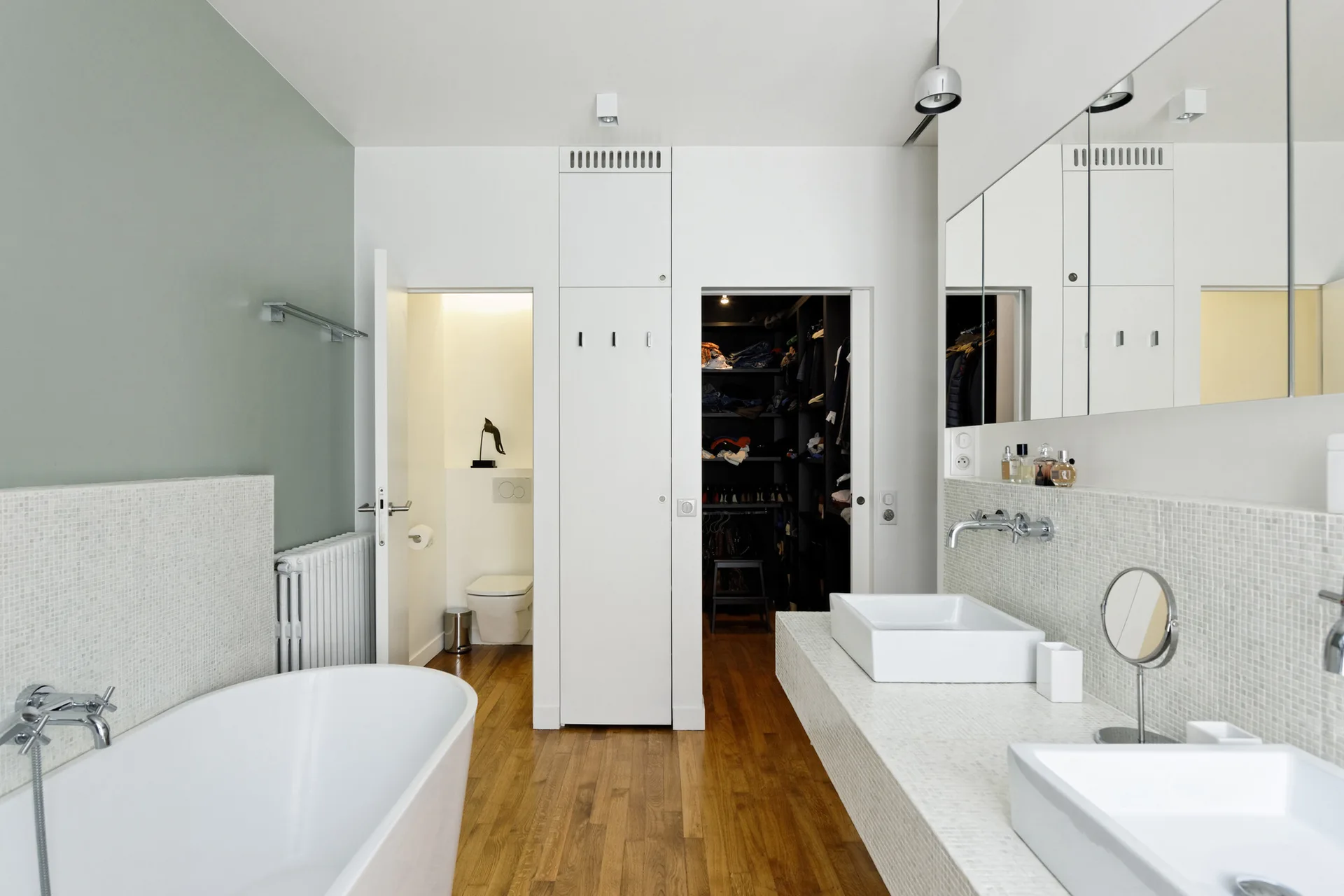 A modern bathroom featuring a white freestanding bathtub, a double vanity covered in small mosaic tiles, and warm wood flooring.