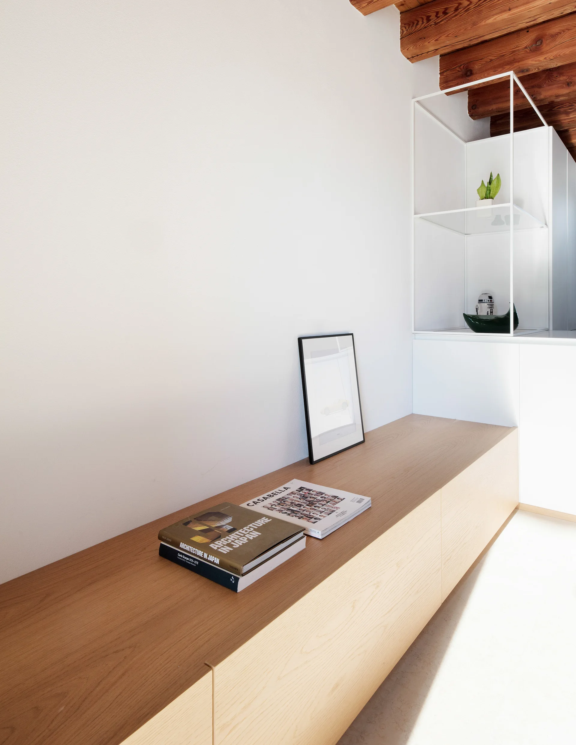 A low oak storage bench sits against a blank white wall below exposed dark wood ceiling beams next to a minimalist white metal shelving unit.