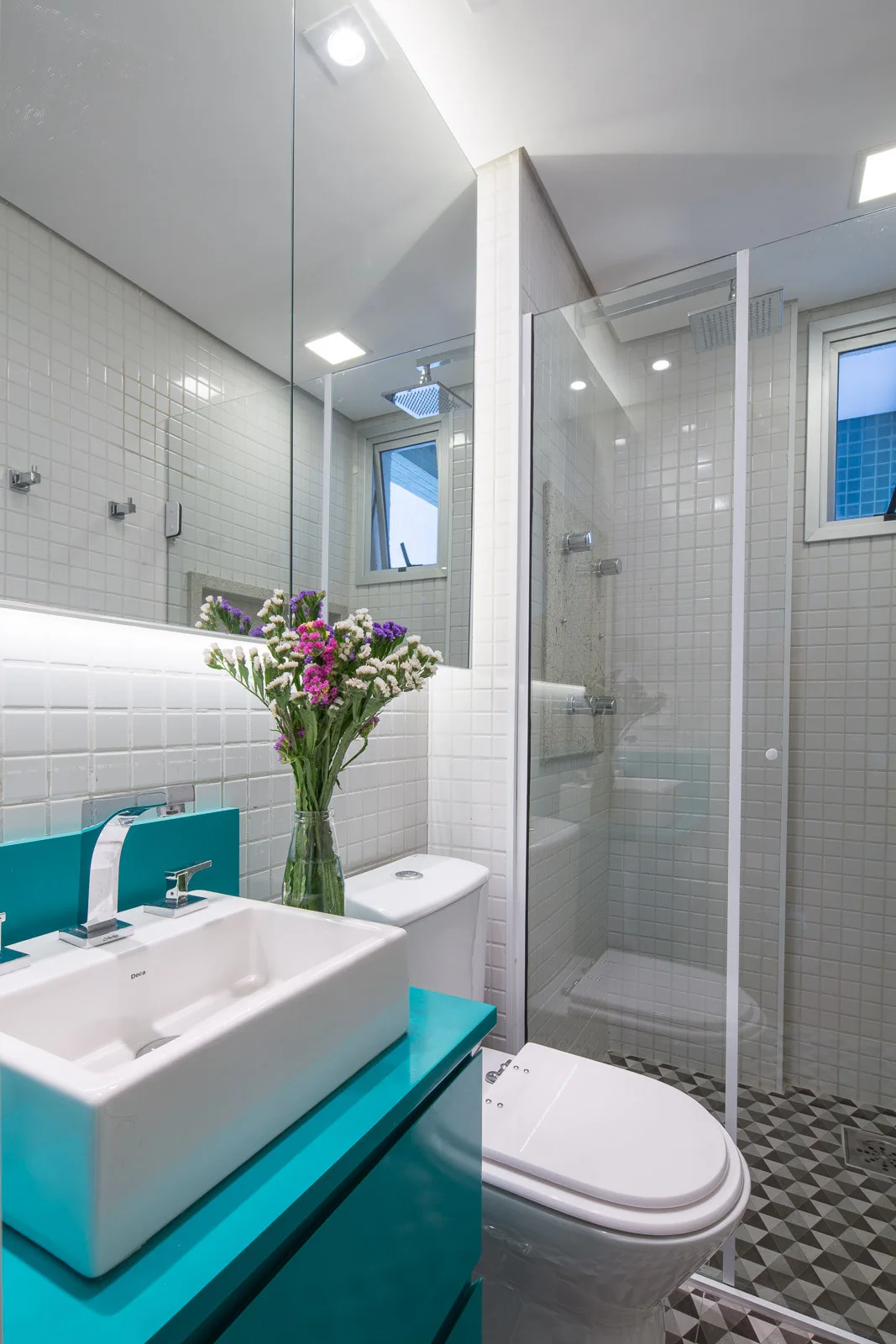 A contemporary bathroom features a bright turquoise vanity with a vessel sink, white square-tiled walls, and a glass-enclosed shower.