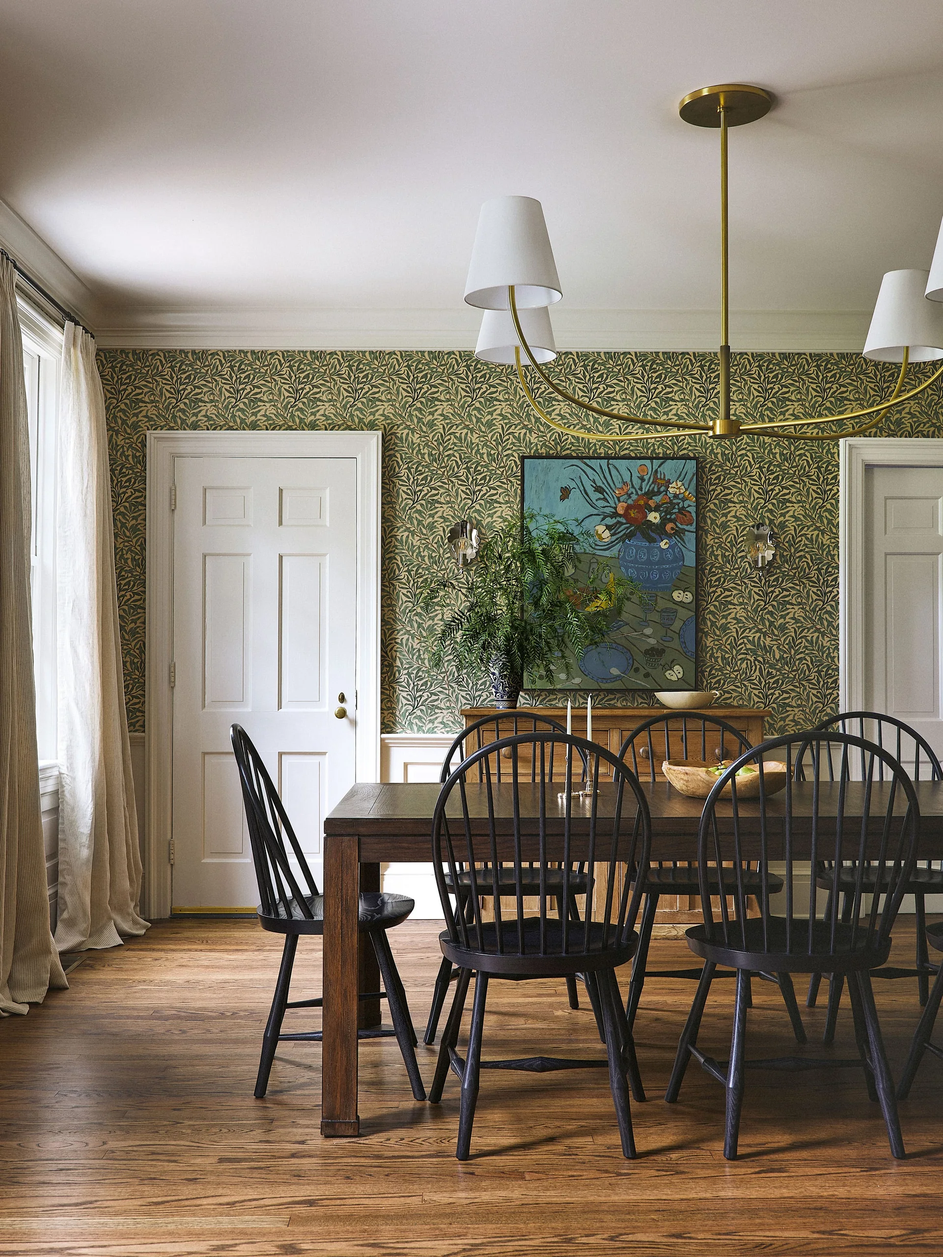 A traditional dining room featuring a wood table, black spindle chairs, green botanical wallpaper, and white wainscoting.