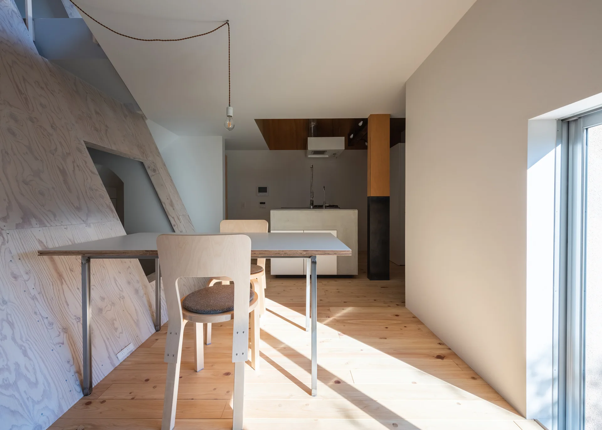 A minimalist dining area featuring a light wood table and chairs next to a slanted plywood wall with a kitchen island in the background.