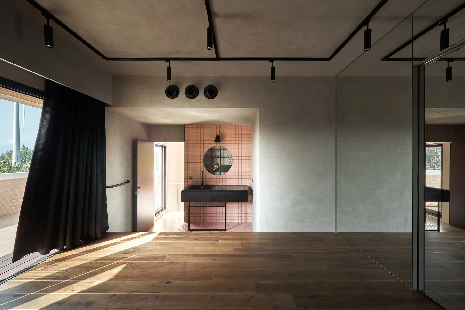 A black floating sink vanity stands in a nook lined with pink square tiles, adjacent to a room with wood flooring and concrete-look walls.