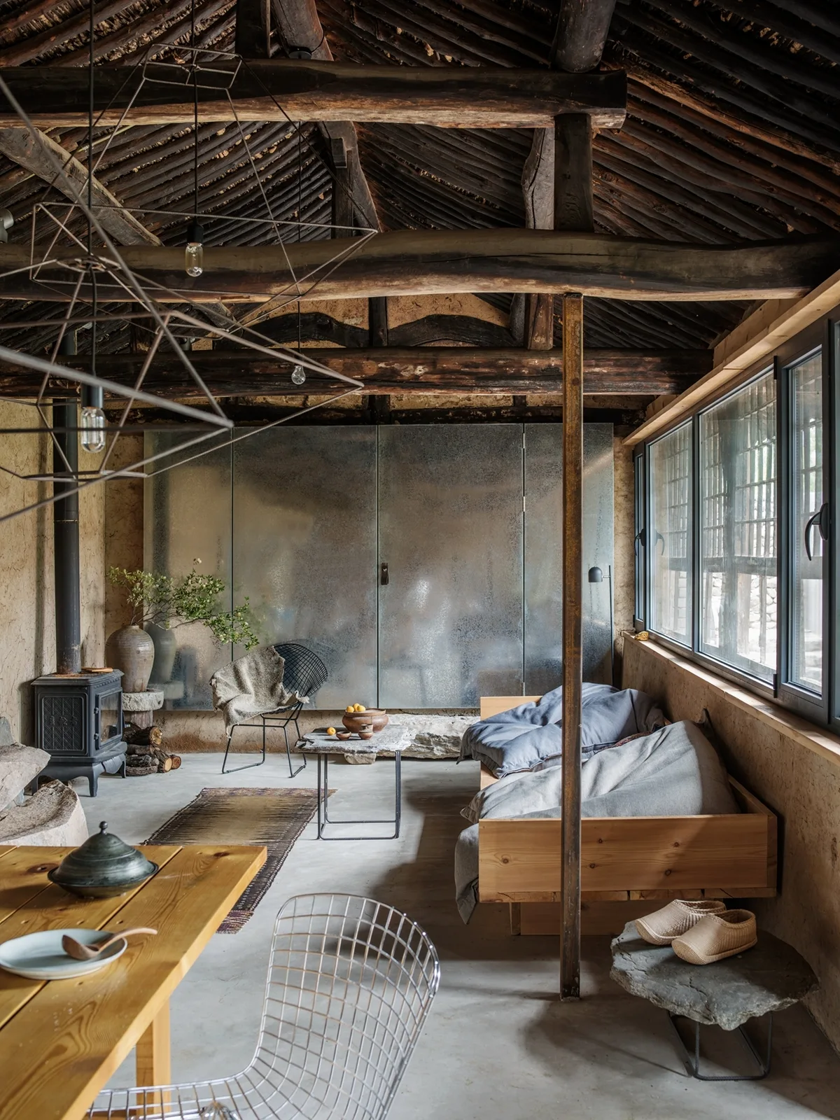 A rustic living room features dark wood log rafters, a wooden daybed under a long window, concrete floors, and a frosted metal partition.