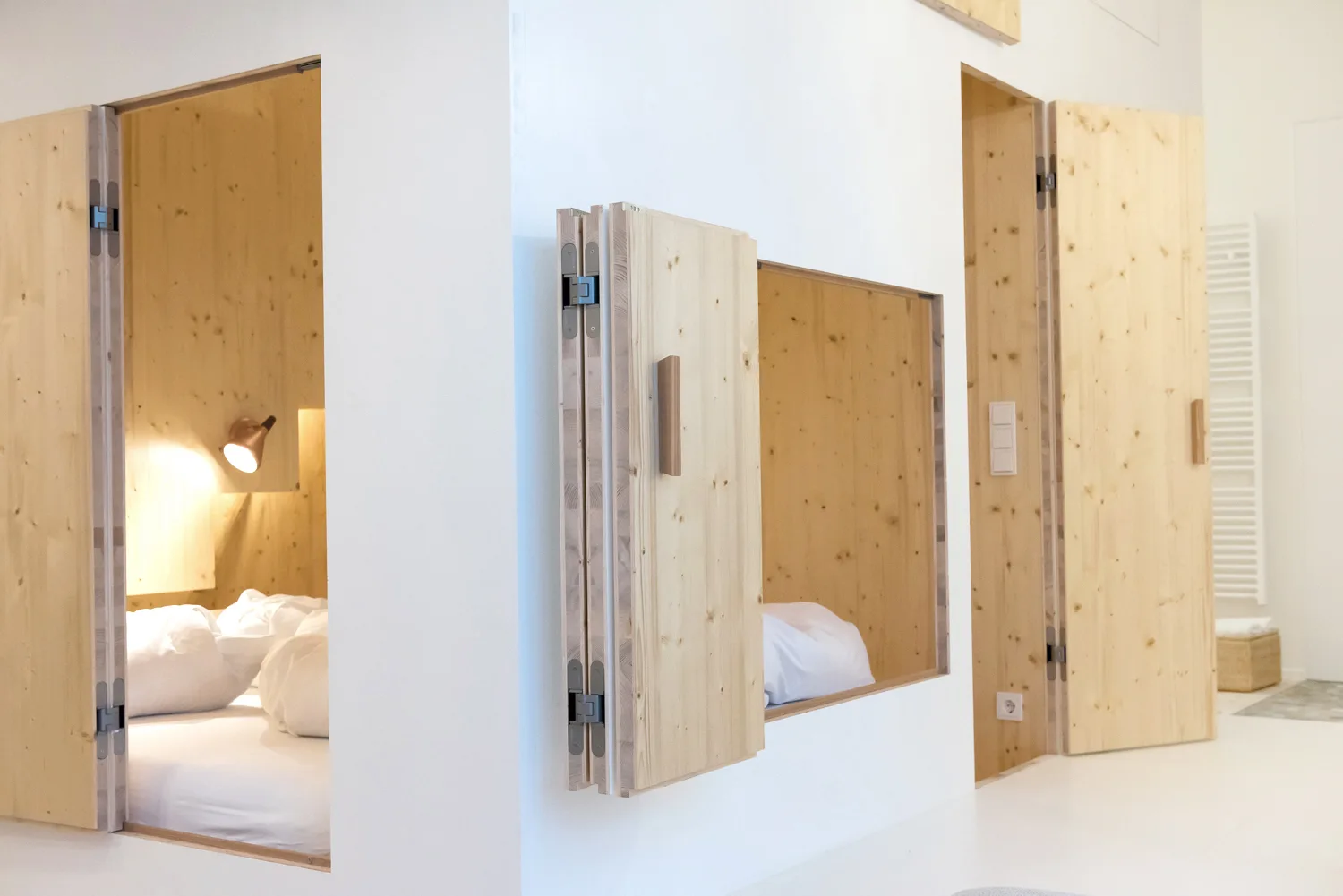 Built-in light wood sleeping alcoves with folding doors set into smooth white walls, featuring white bedding and a warm interior copper sconce.