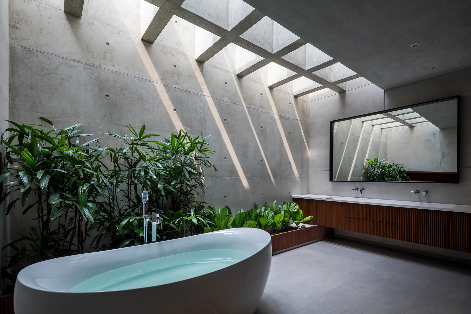 A modern bathroom features a freestanding white bathtub, a slatted wooden vanity, and green plants set against concrete walls under a grid skylight.