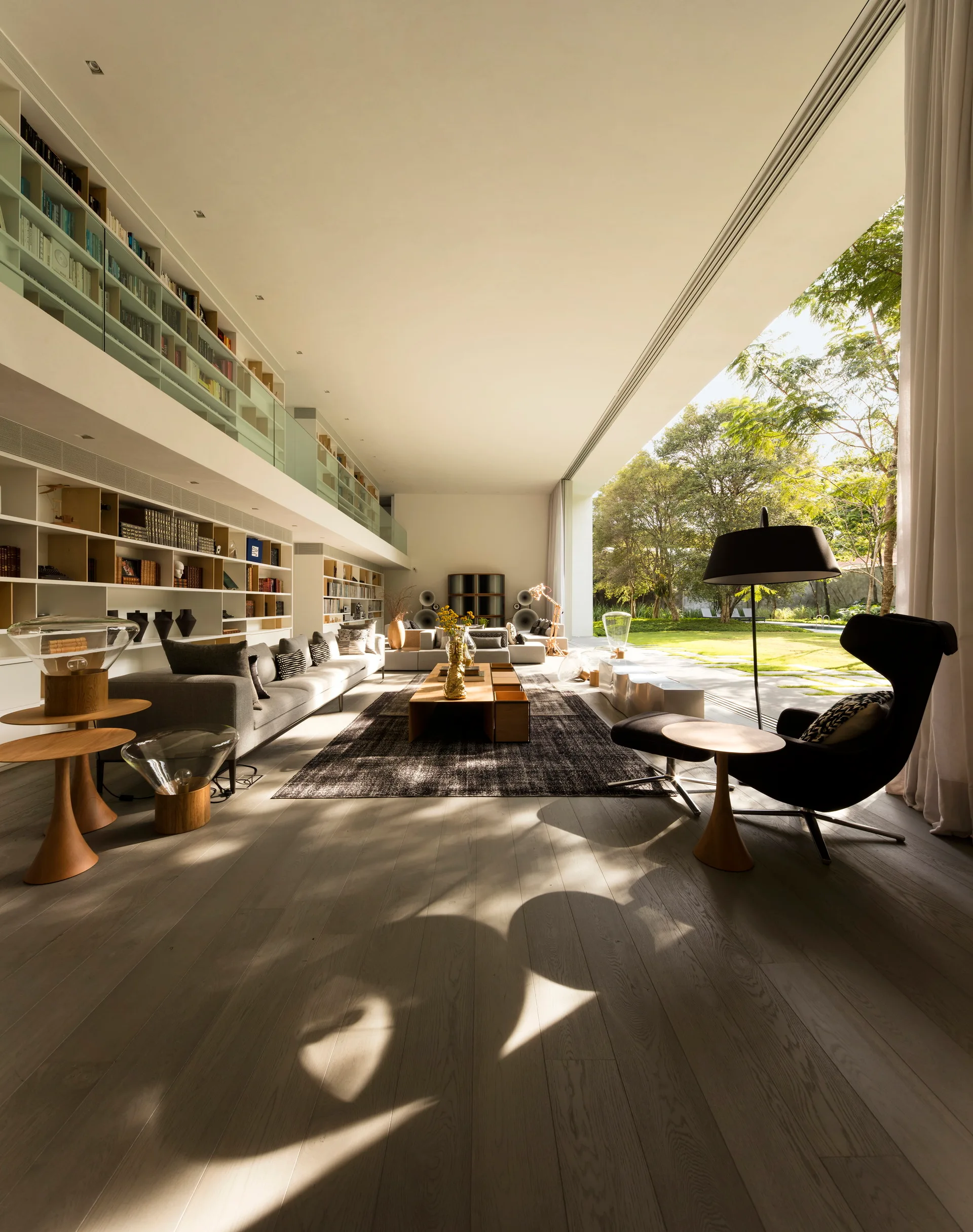 A spacious double-height living room features a multi-level built-in library, a low grey sectional sofa, and a glass wall opening to a green garden.