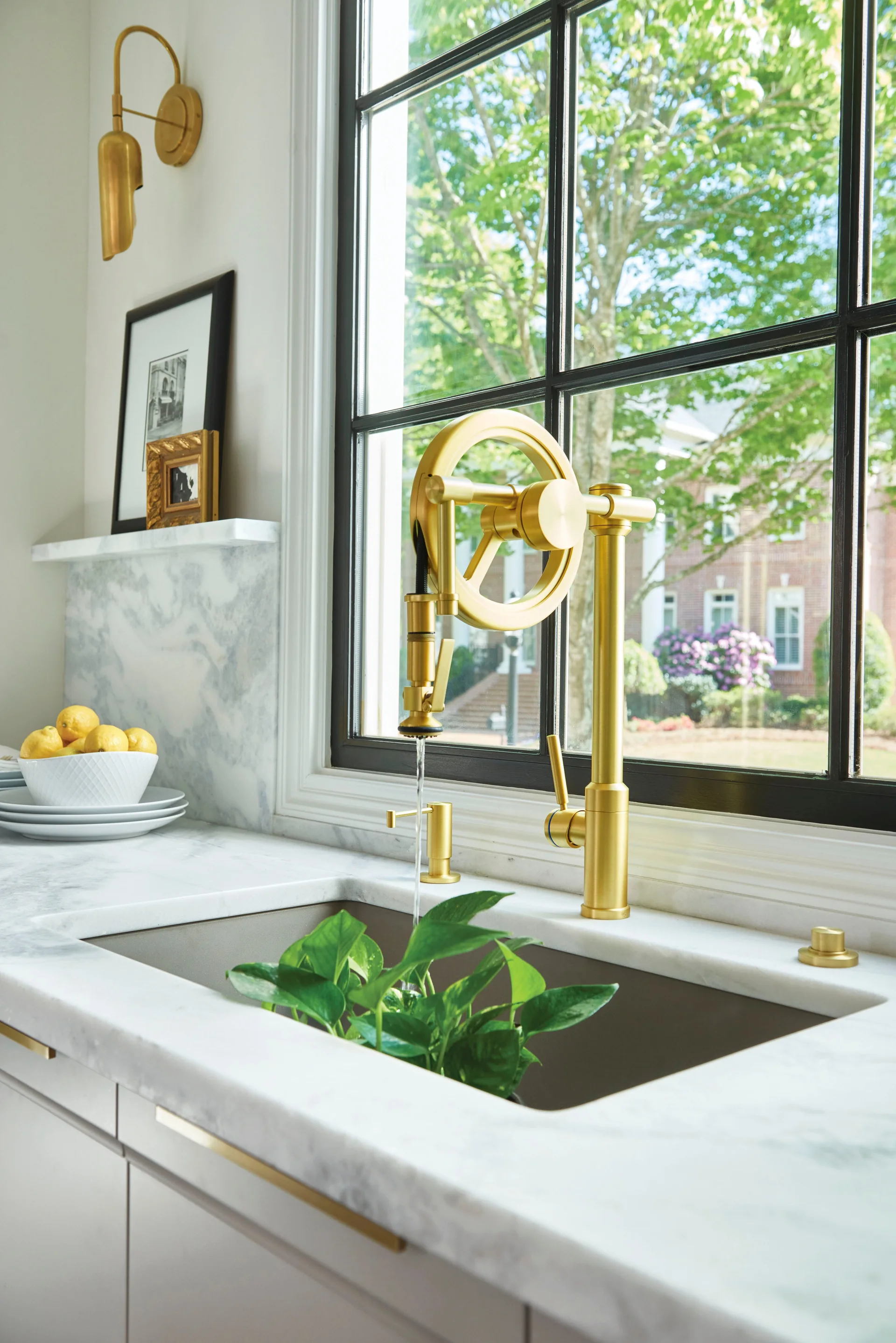 A gold-toned industrial wheel faucet is mounted on a white marble countertop under a black-framed window, next to a matching brass sconce.