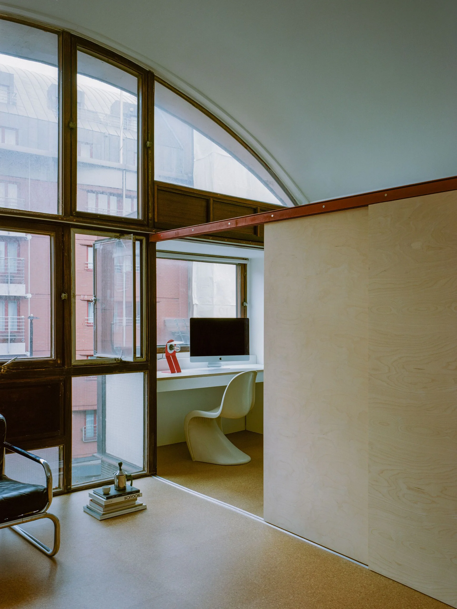 A minimalist home office features a white built-in desk, a sculptural white chair, and a light wood sliding partition beside large gridded windows.