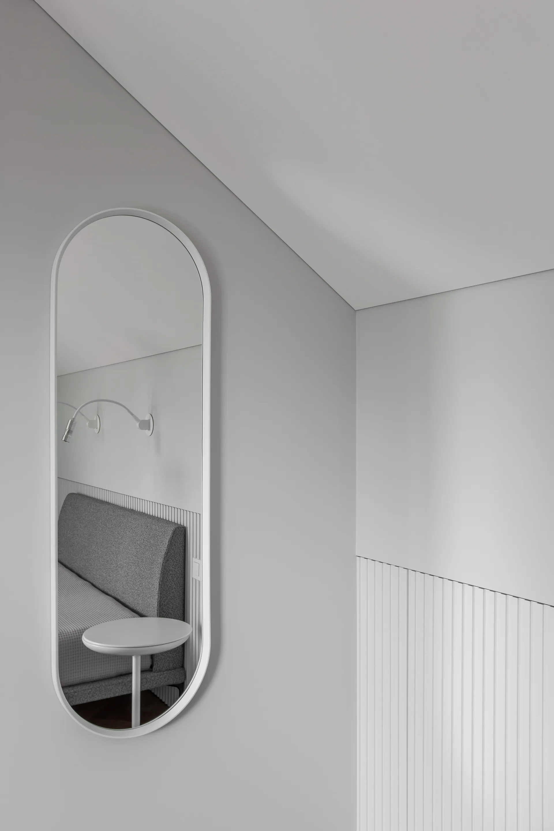A tall oval mirror with a white frame hangs on a grey wall above vertical wood slats, reflecting a grey headboard and small white nightstand.