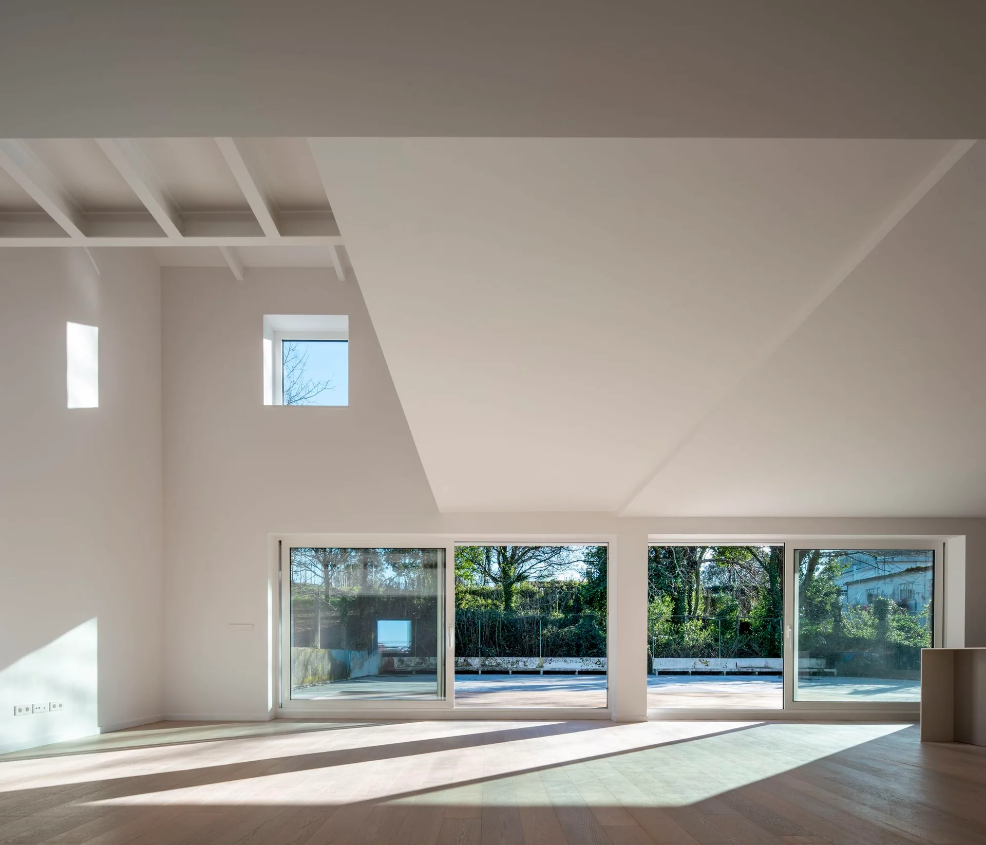 A spacious minimalist room featuring light wood flooring, plain white walls, exposed ceiling rafters, and large sliding glass doors leading outside.