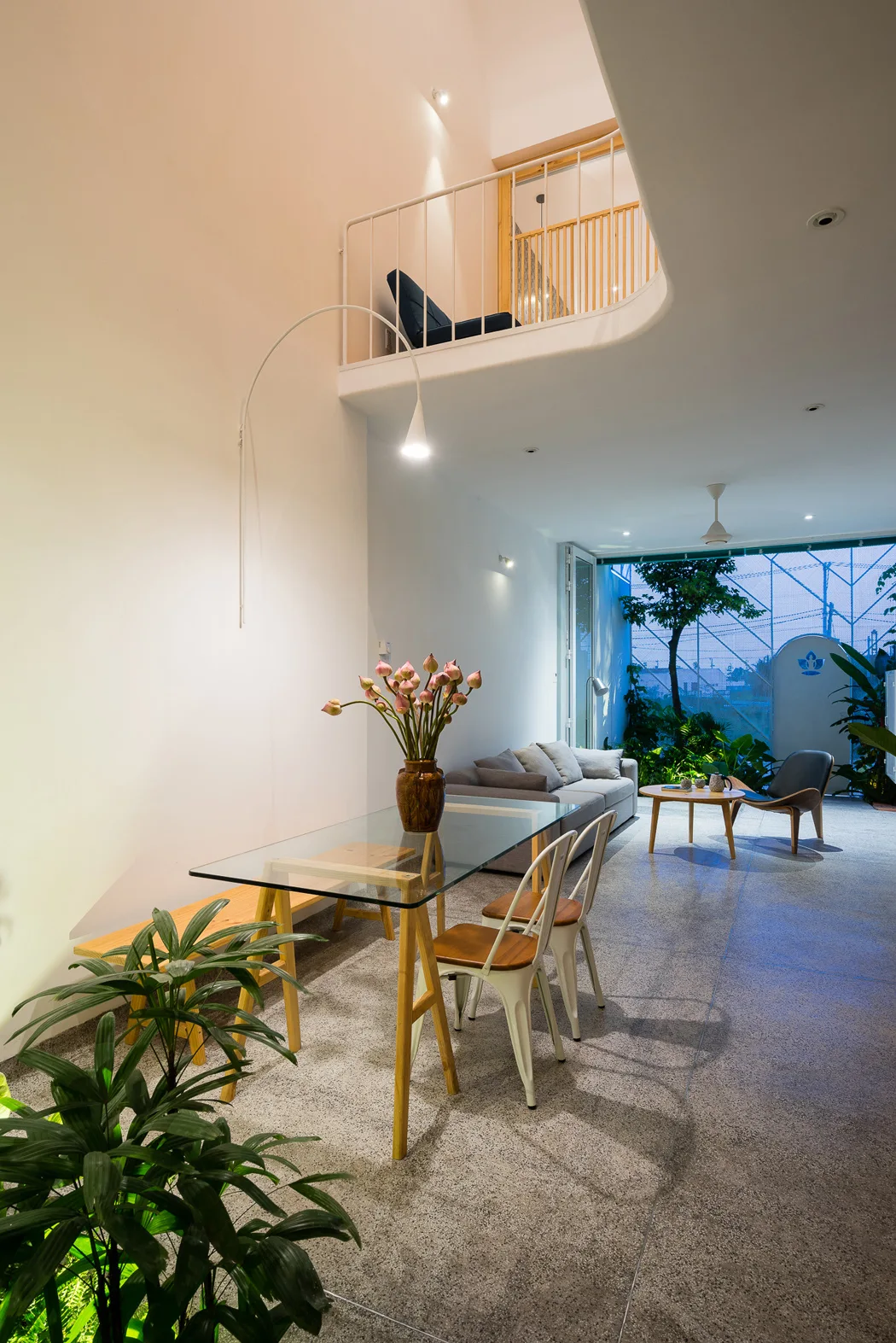 A modern open-plan dining and living area features a glass-top table, speckled grey flooring, a grey sofa, and a white mezzanine balcony overhead.