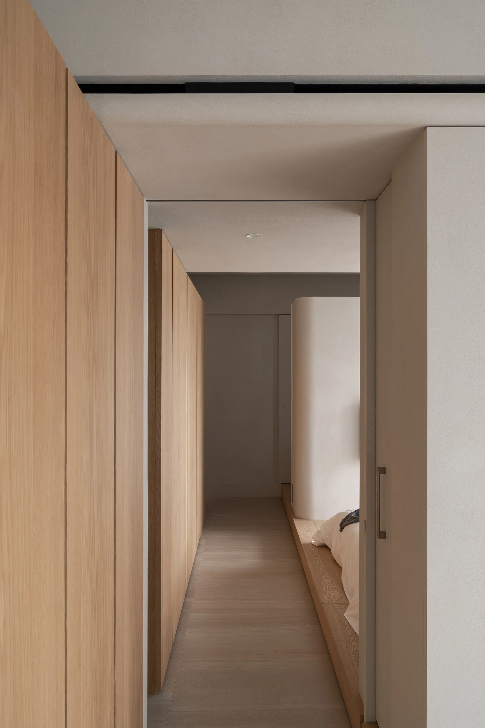 A narrow hallway featuring floor-to-ceiling light wood cabinetry leads into a minimalist bedroom with a low platform bed and curved white wall.