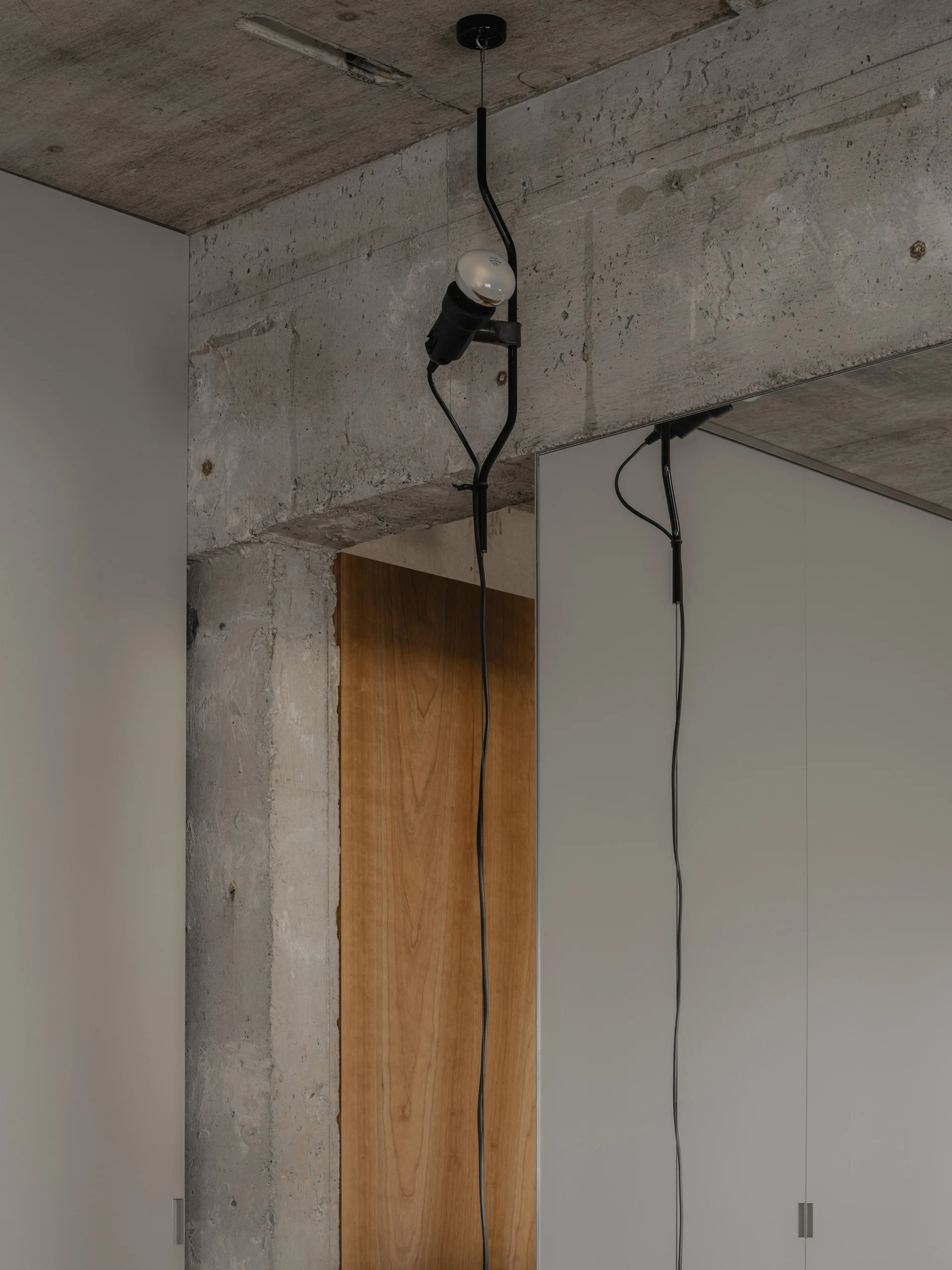 A black industrial pendant light hangs near an exposed concrete ceiling and column next to matte grey cabinet panels and warm wood paneling.