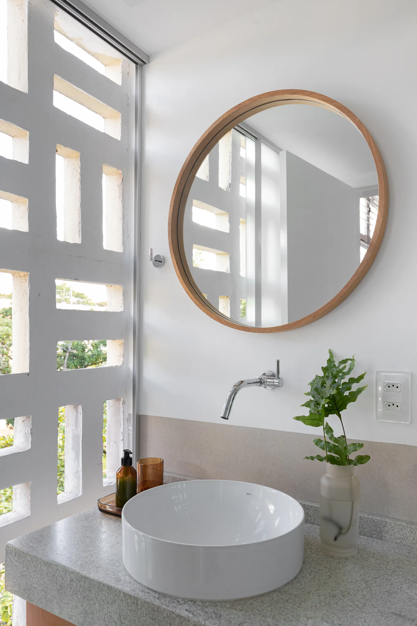 A modern bathroom vanity featuring a round white vessel sink, a wall-mounted faucet, and a round wood-framed mirror next to a white breeze block wall.