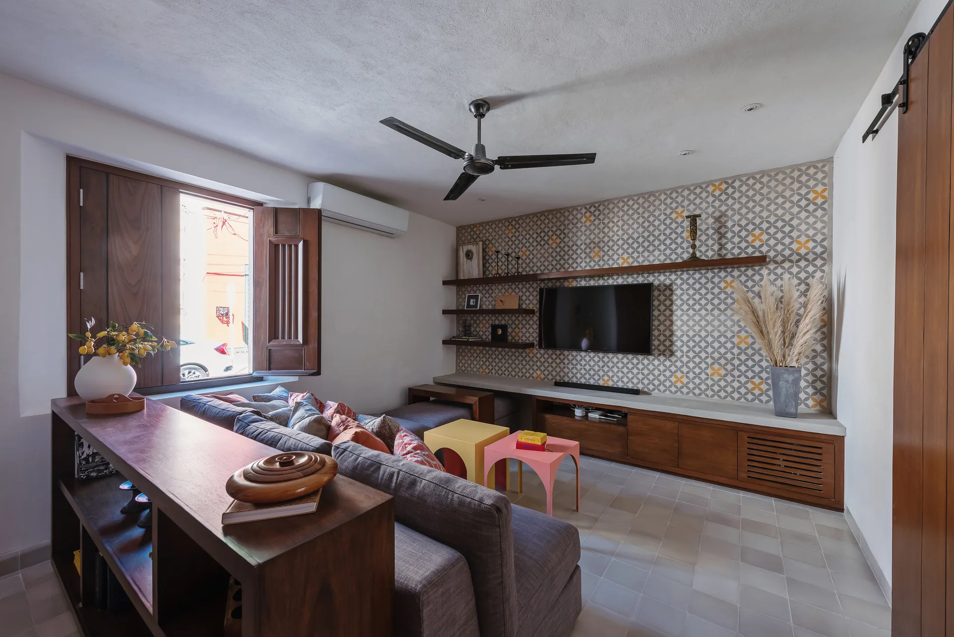 A modern living room featuring a grey sectional sofa, wooden console, geometric patterned accent wall with a wall-mounted TV, and wood shutters.