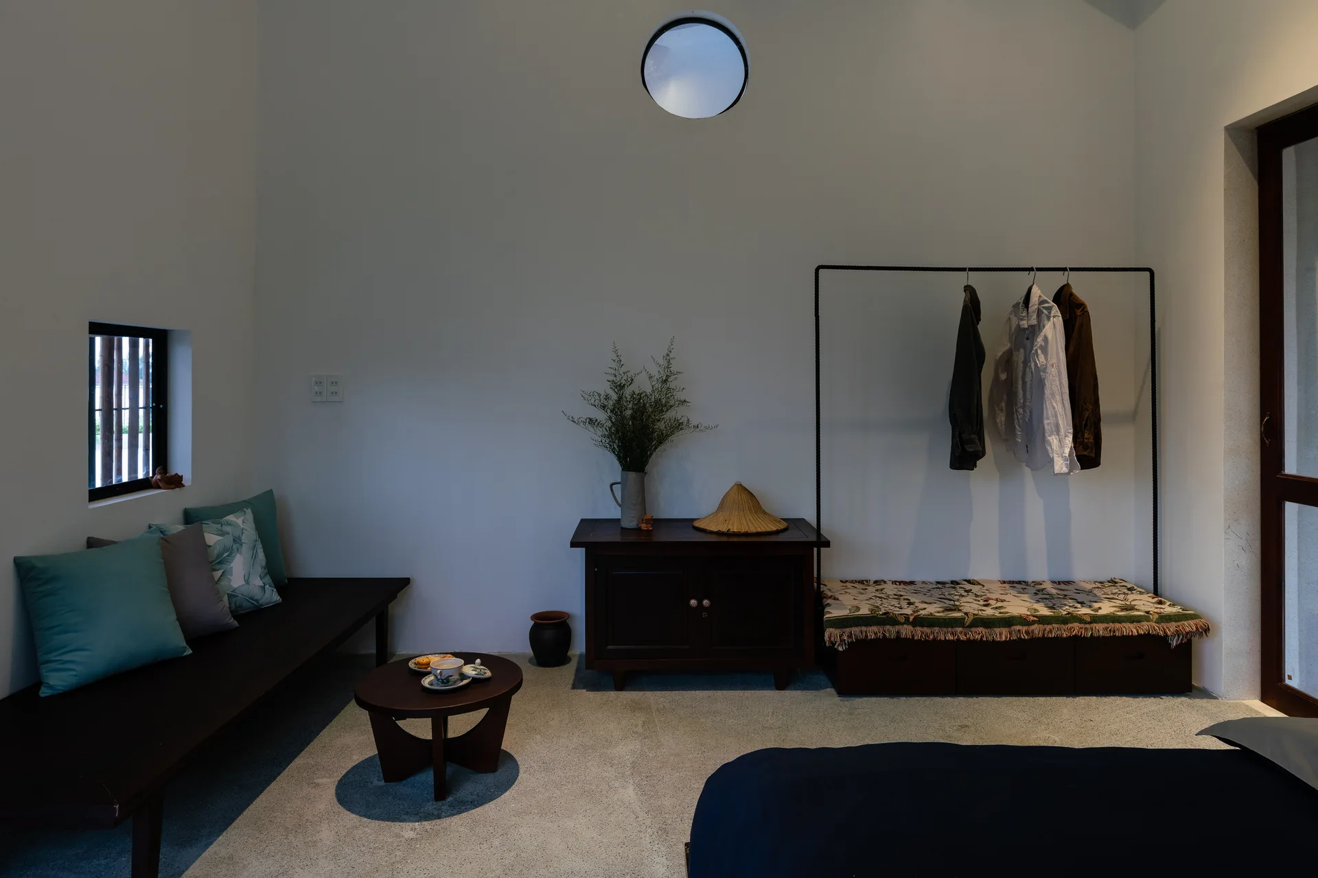 A minimalist bedroom featuring white plaster walls, a circular oculus window, dark wooden benches, and light grey concrete flooring.