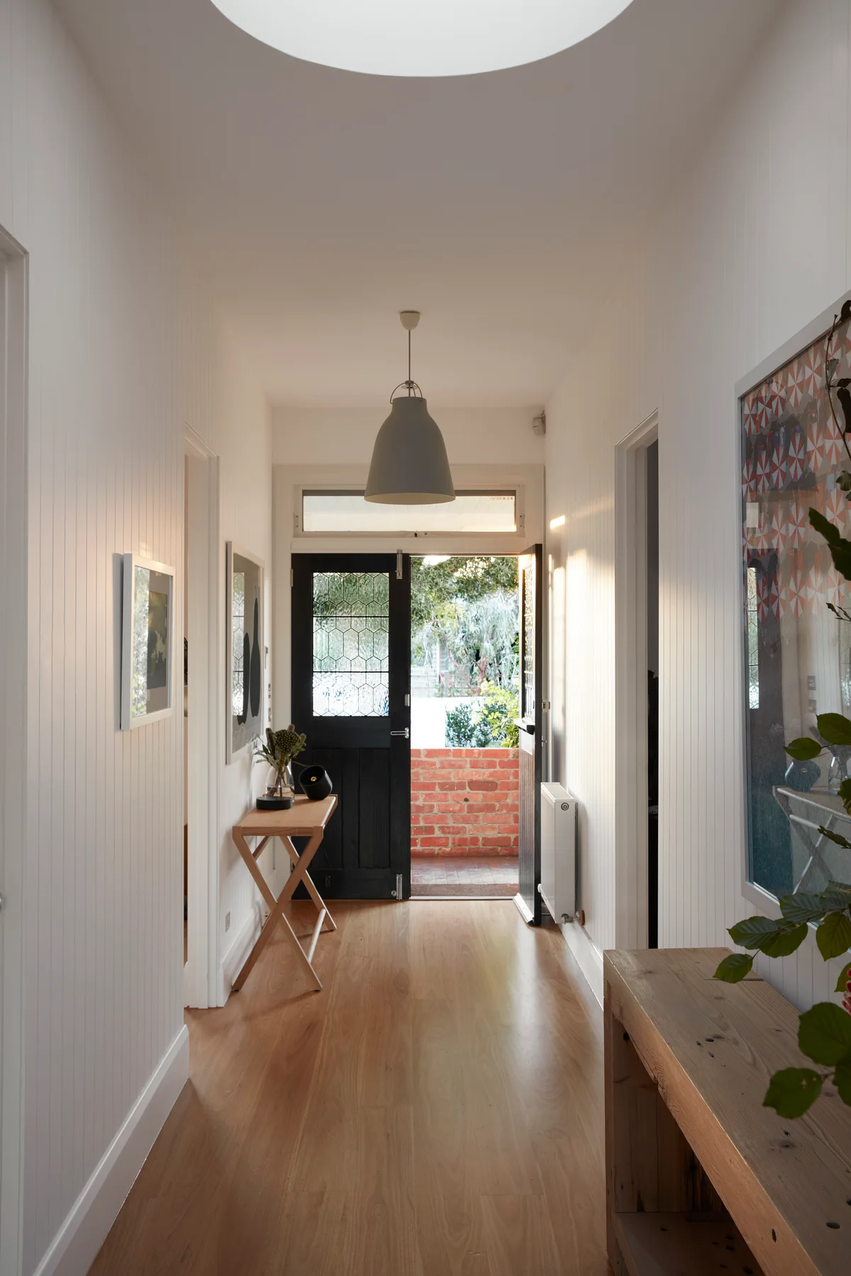 A bright white hallway featuring vertical wood wall paneling, light wood flooring, a black entry door, and a circular skylight above.