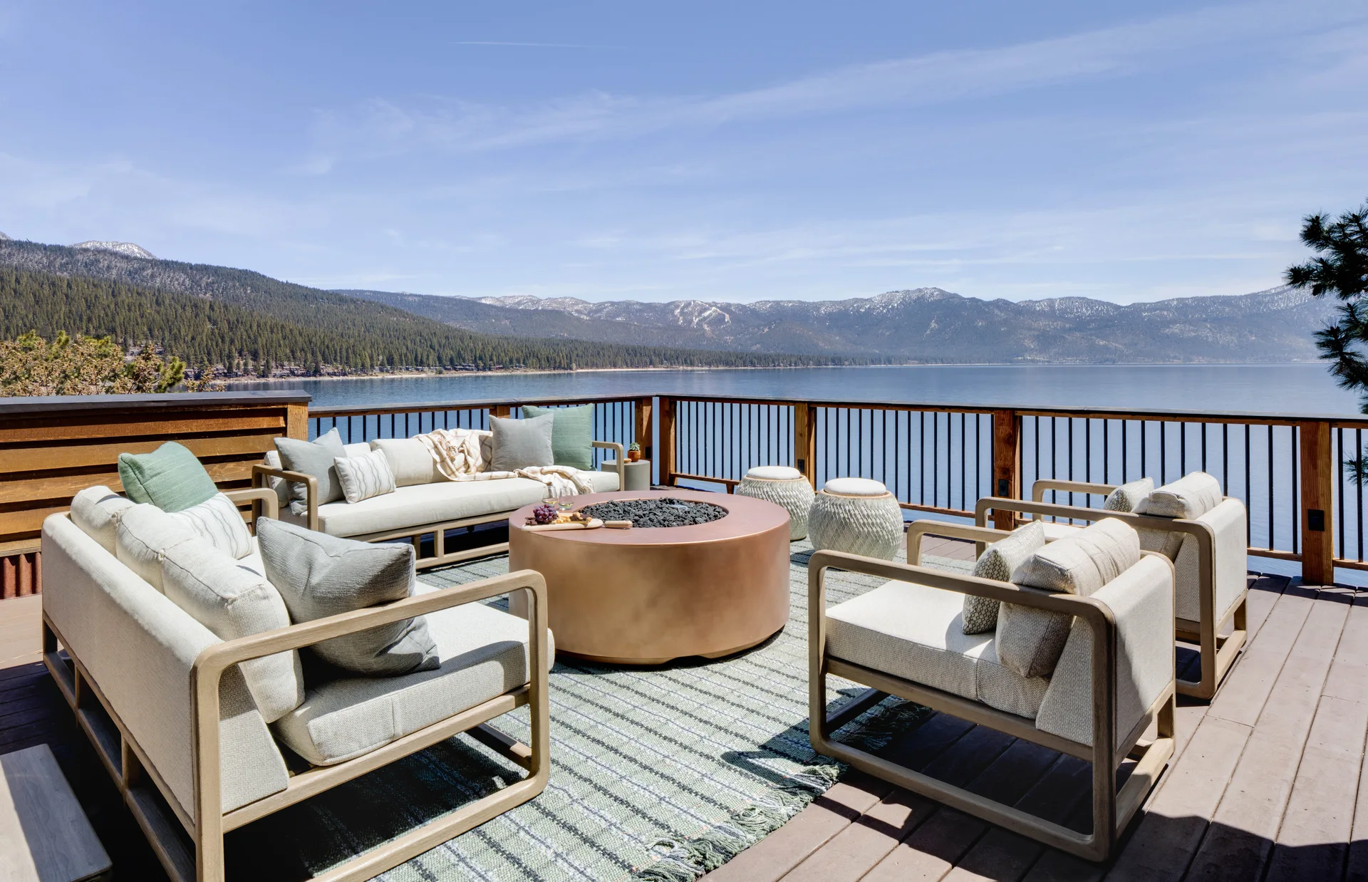 Modern neutral-toned outdoor sofas and armchairs are arranged around a circular copper fire pit on a wooden deck overlooking a lake and mountains.