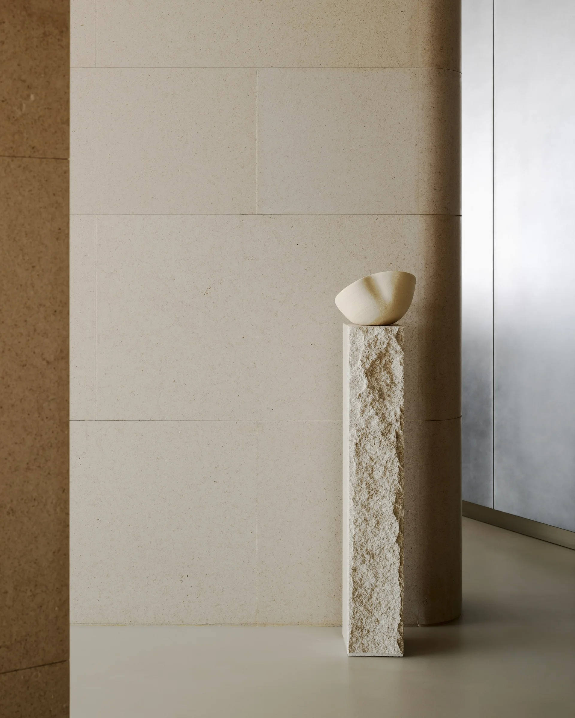 An organic cream ceramic bowl sits atop a tall, rough-textured stone pedestal against smooth beige-paneled walls in a minimalist interior.