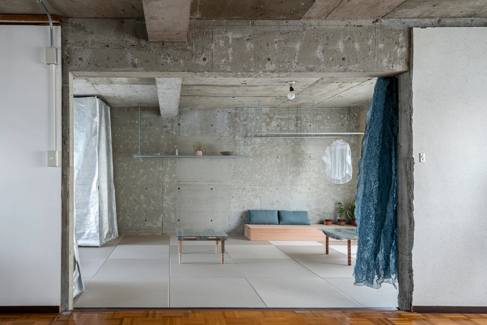 An open minimalist room features raw exposed concrete walls and ceiling, light-colored tatami floor mats, and simple low wooden furniture.