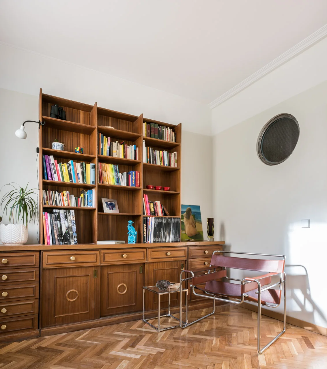 A study corner features a large wooden bookcase filled with books, a brown leather and metal armchair, and patterned herringbone parquet flooring.