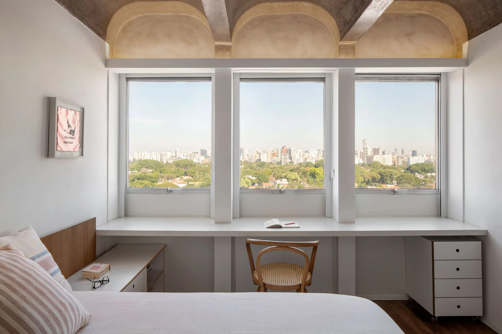A minimalist bedroom features a long white desk built under a triple window, a wooden chair, and an exposed concrete vaulted ceiling.