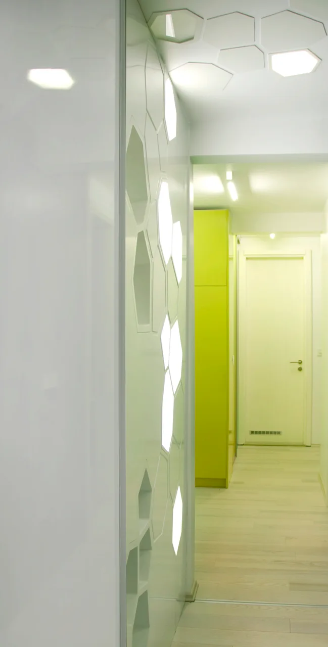 A modern white hallway features a glossy wall with backlit hexagonal panels, a light wood floor, and a lime green built-in cabinet.