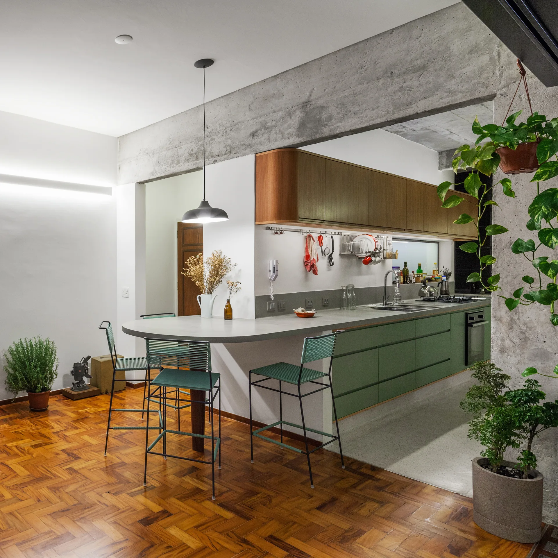 A modern kitchen featuring sage green lower cabinets, a curved grey concrete breakfast bar with green stools, and warm wood herringbone flooring.