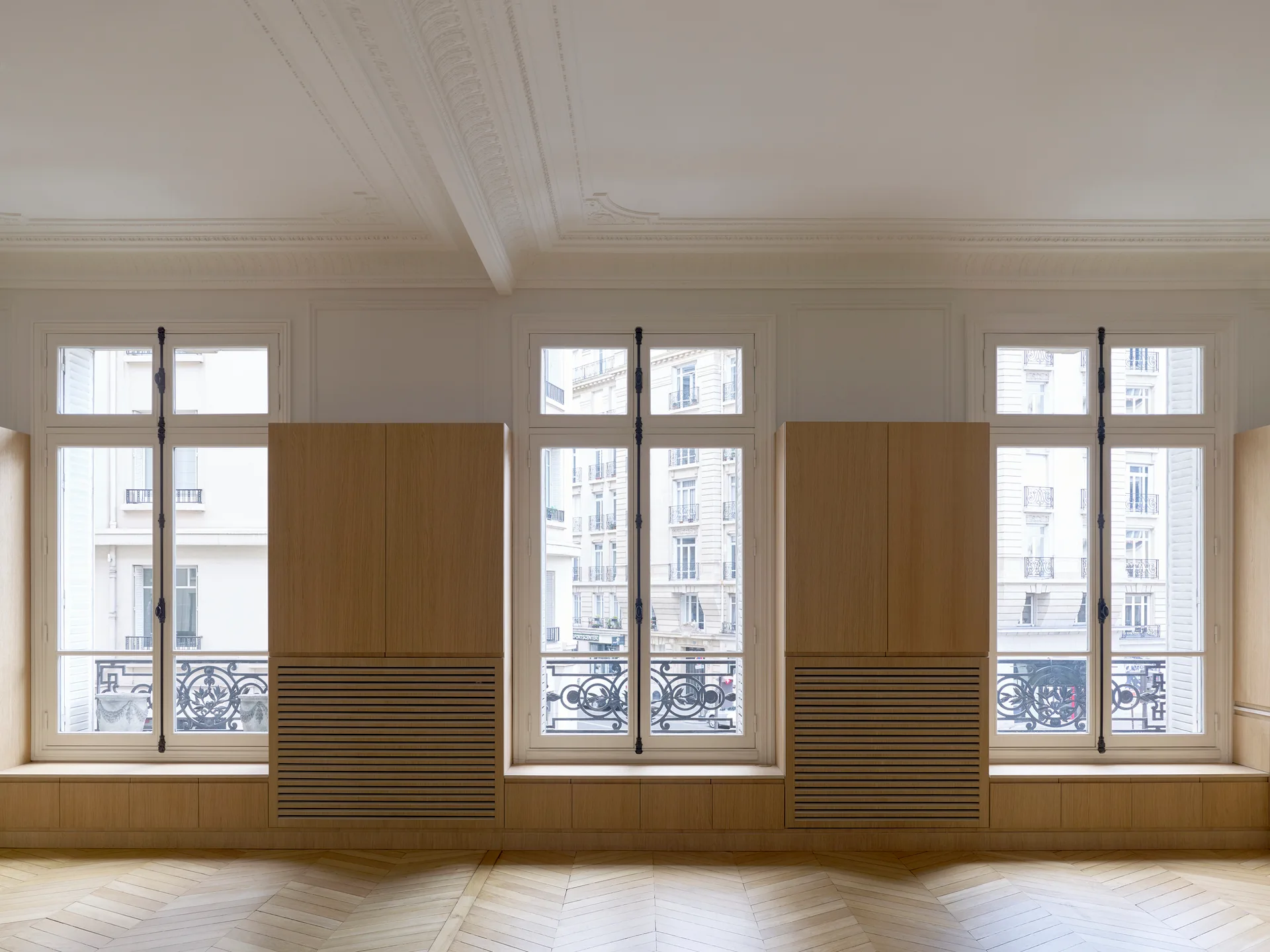 An airy room features custom oak built-in cabinets with slatted lower panels set between tall French windows above herringbone parquet wood flooring.