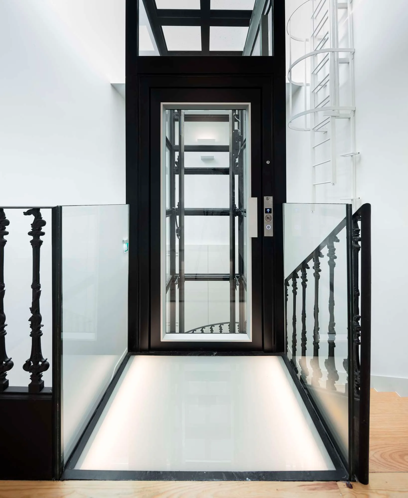 A modern glass elevator with a black metal frame stands at a landing featuring an illuminated glass floor and decorative dark metal railings.