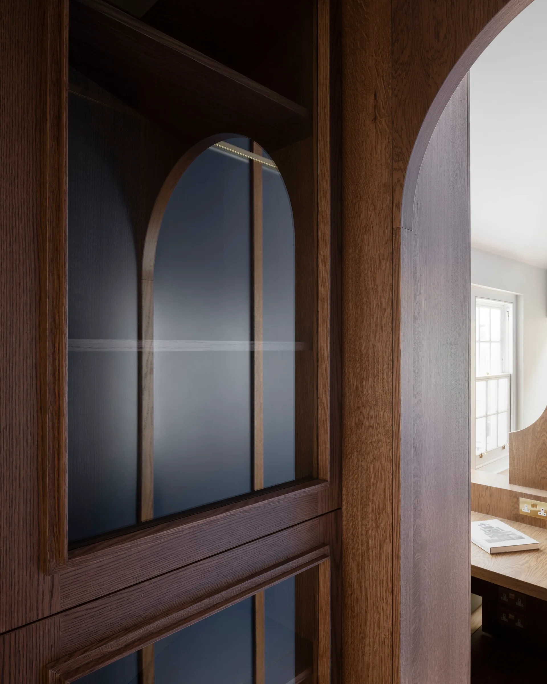 Custom dark wood built-in cabinetry featuring arched glass panels and an adjacent arched doorway opening to a home office desk.