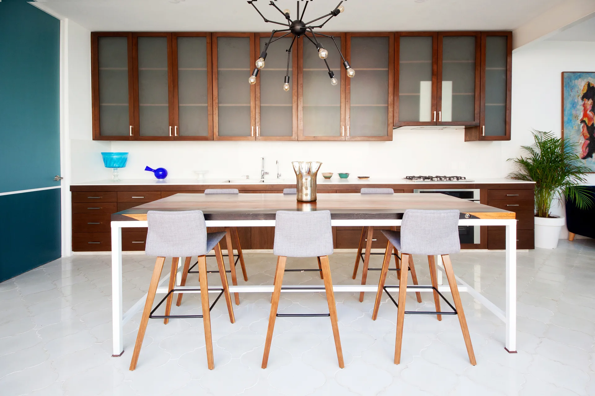 A modern kitchen with dark wood upper and lower cabinets, a central wood-topped dining island with gray stools, and white geometric floor tiles.