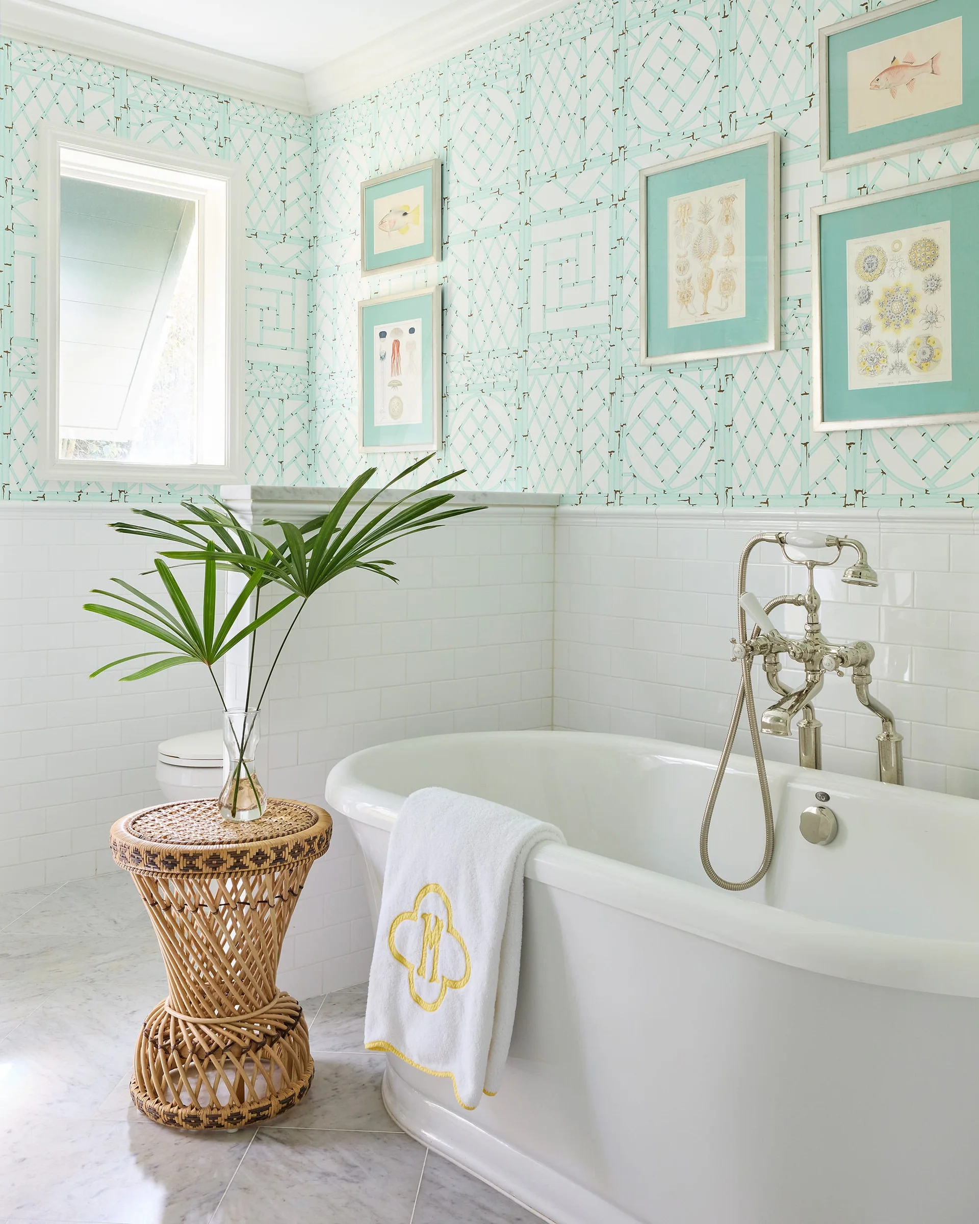 A white freestanding bathtub stands in a bathroom featuring light blue lattice wallpaper, white subway wainscoting, and a woven rattan side table.