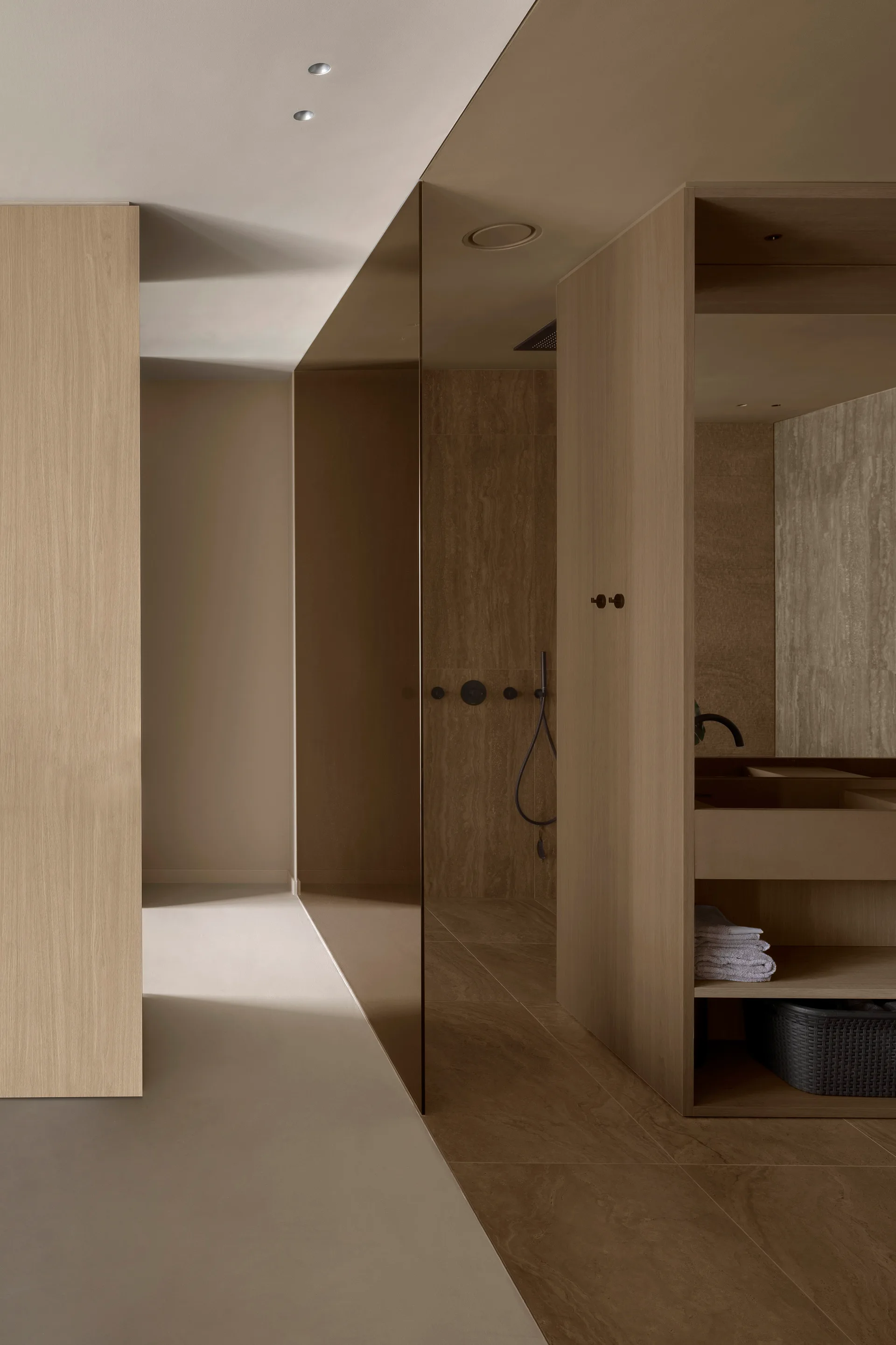 A minimalist bathroom features wood-toned paneling, stone tiles, and a bronze tinted glass partition separating the vanity from the shower area.