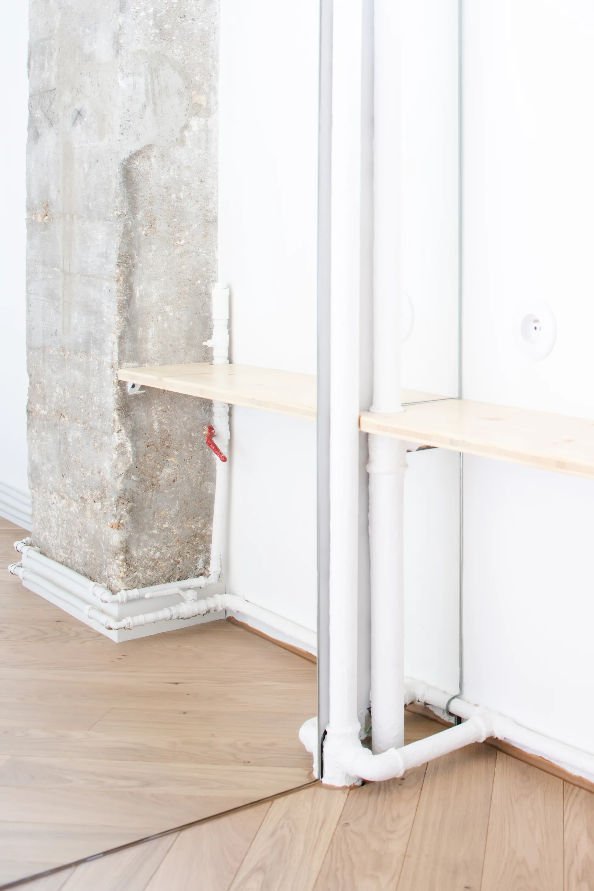 A light wood shelf mounted next to a rough concrete column and white walls with painted pipes reflected in a wall mirror.