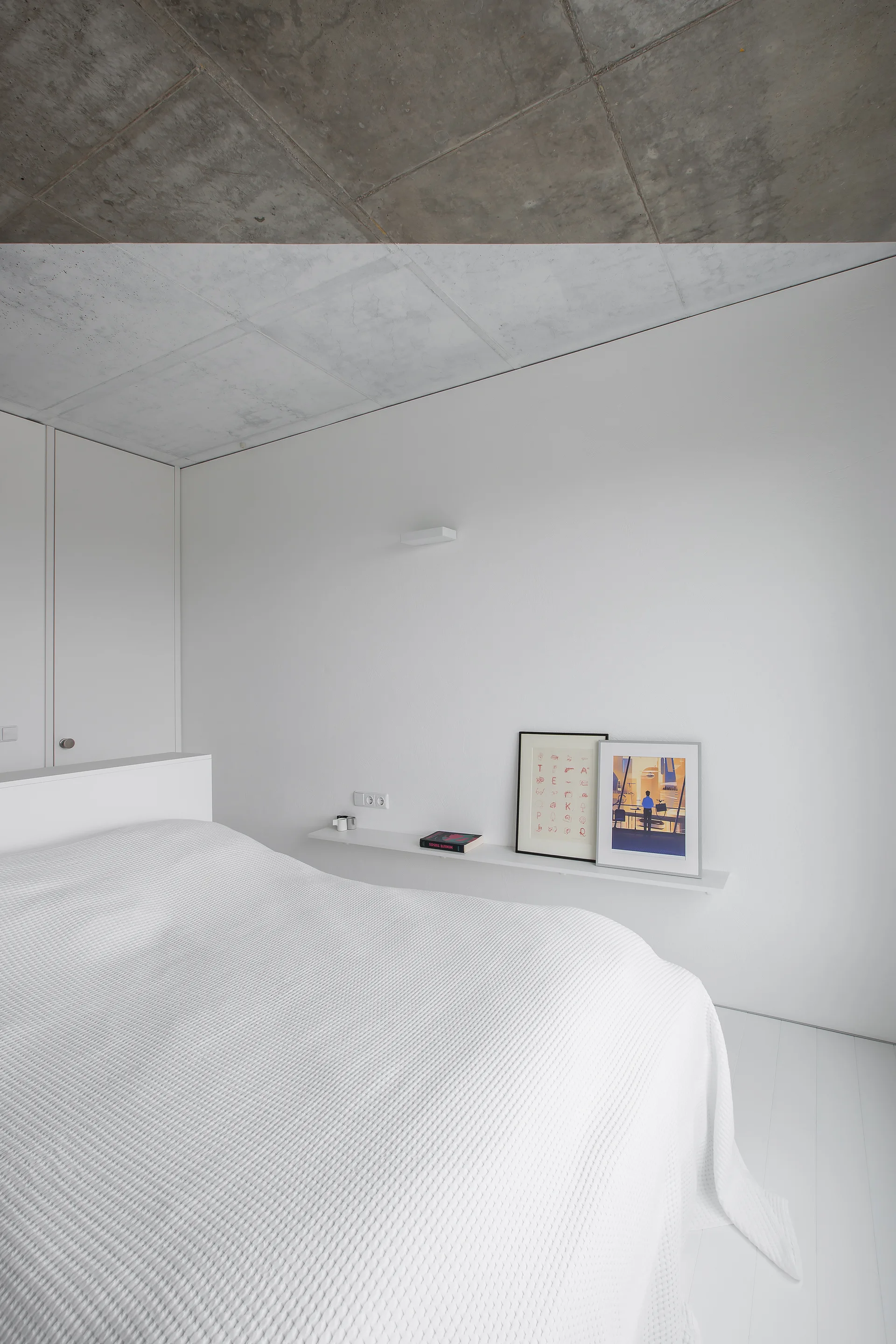 A minimalist white bedroom featuring a bed with waffle-textured bedding, a long floating shelf with art frames, and an exposed concrete ceiling.