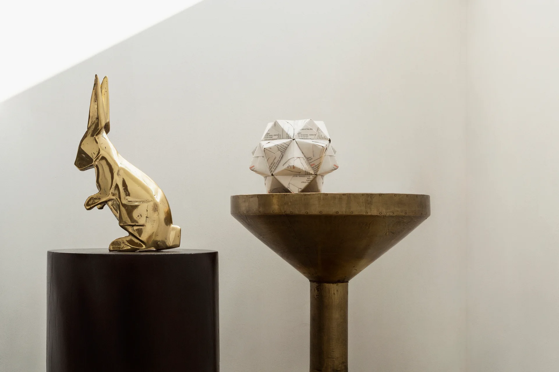 A brass rabbit sculpture and a folded paper geometric sphere rest on metal pedestals against a white wall with diagonal sunlight.