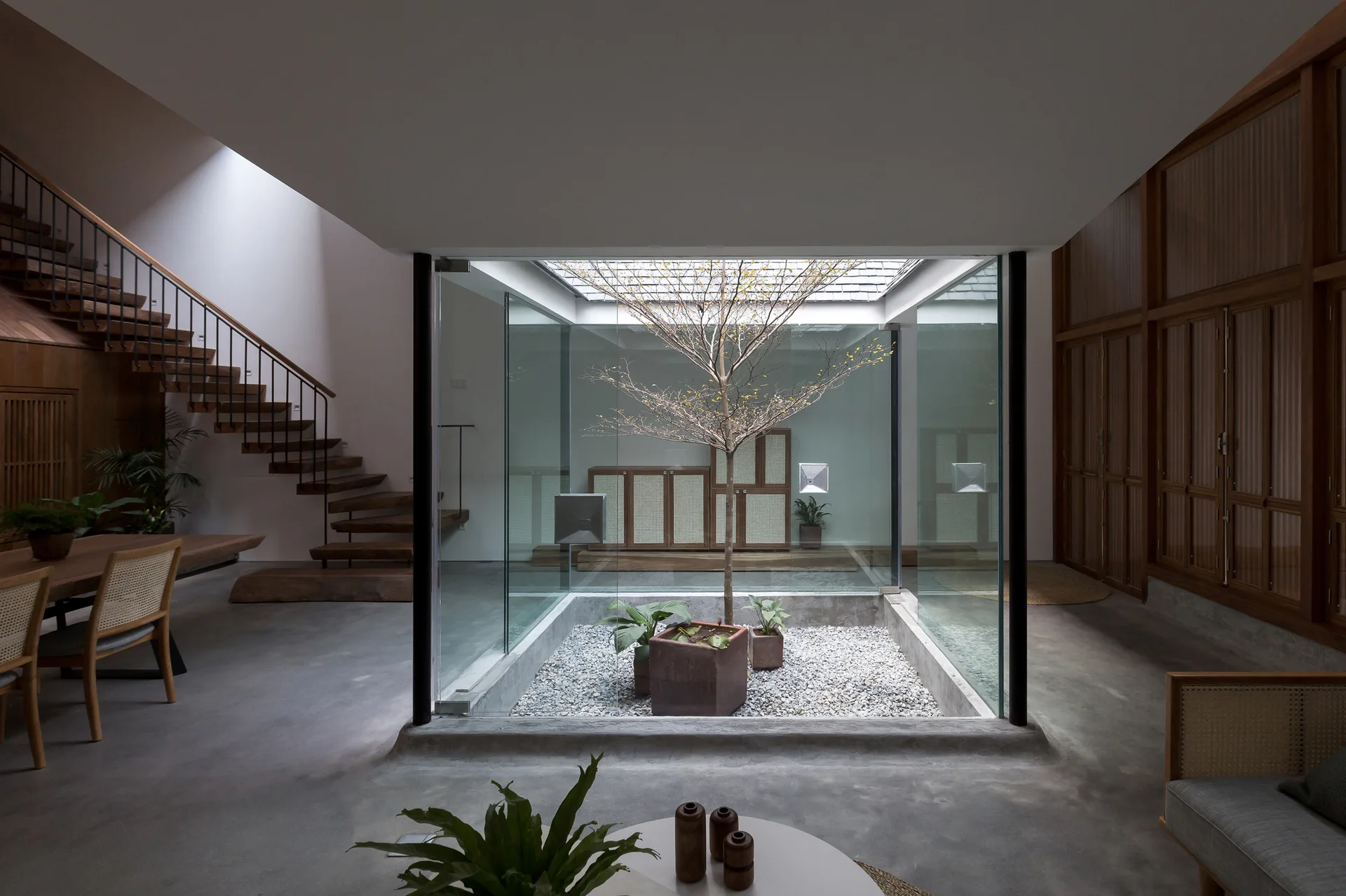 A modern living space featuring a central glass-enclosed garden with a small tree, surrounded by concrete flooring and a wooden staircase.