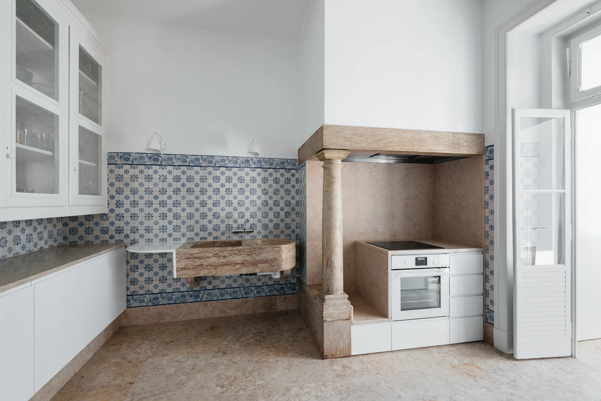 A kitchen features a carved stone sink and a stone oven surround supported by a column against a wall of blue and white patterned tiles.